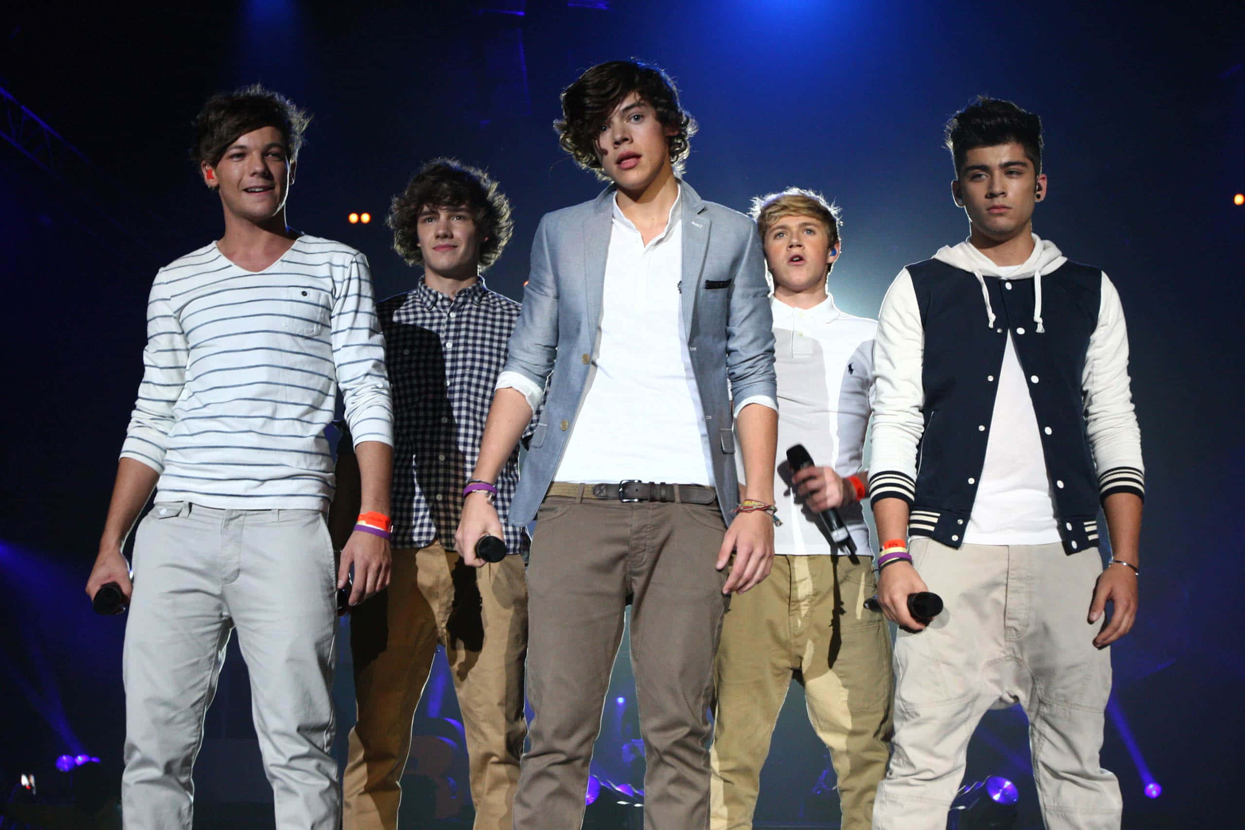 One Direction 桌布 壁紙 — 五位年輕男歌手站在燈光明亮的舞台上，在彩色聚光燈下手持麥克風。