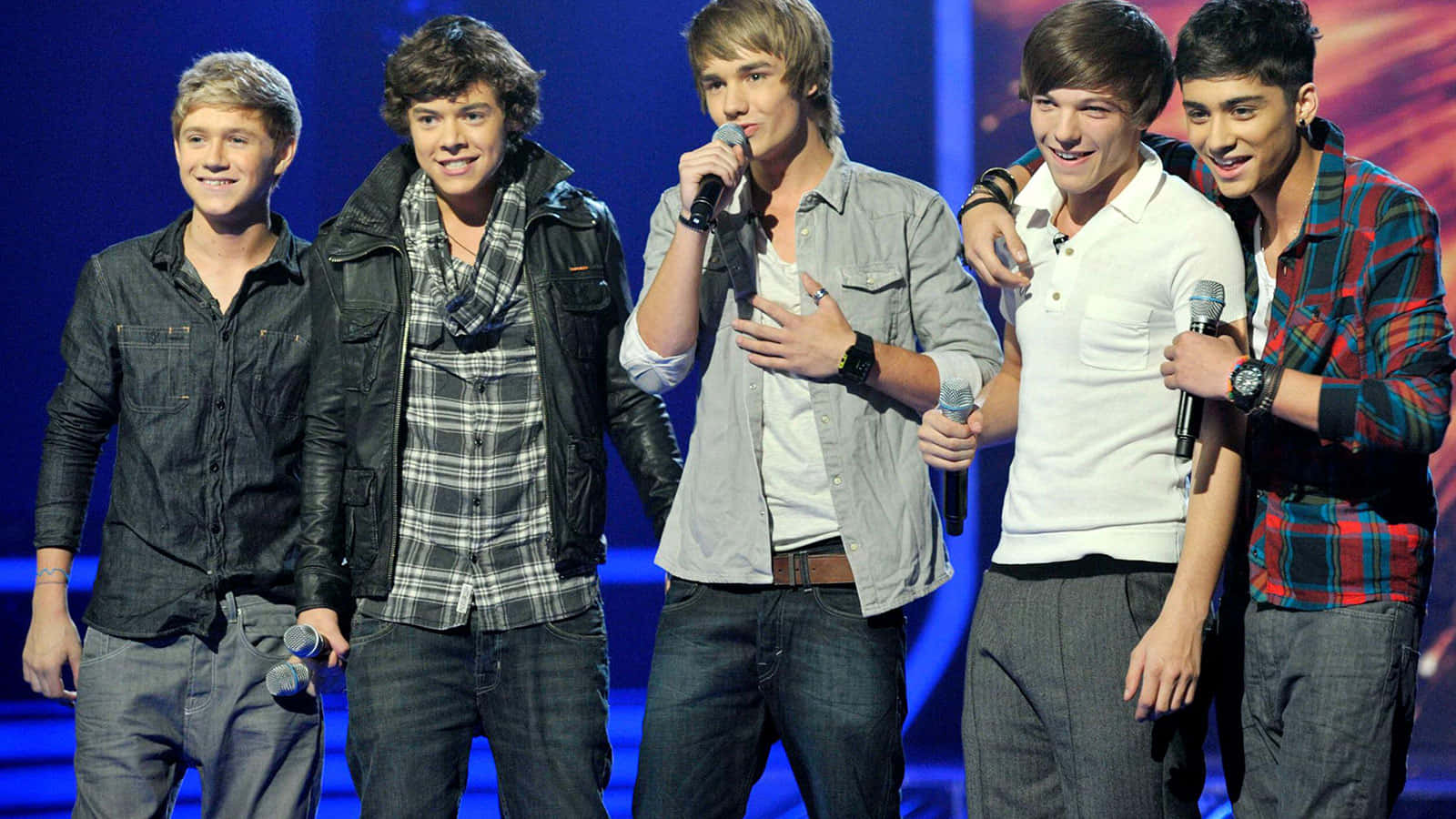 One Direction 桌布 壁紙 — 五位年輕男流行歌手站在燈光明亮的舞台上，手持麥克風，向觀眾微笑。