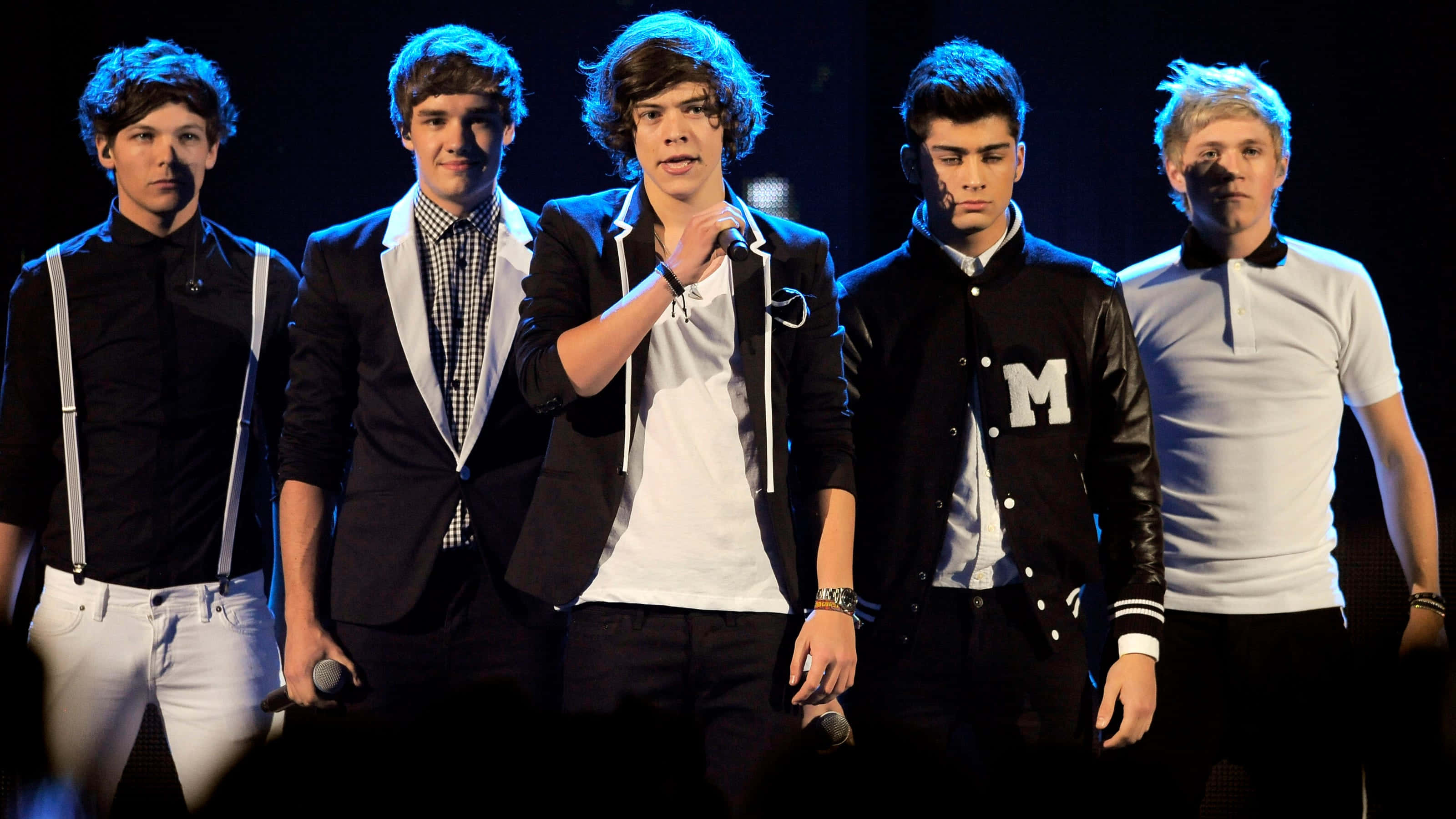 One Direction 桌布 壁紙 — 五位年輕男表演者穿著時尚休閒服裝，站在黑暗的舞台上，在明亮的聚光燈下，其中一人手持麥克風。