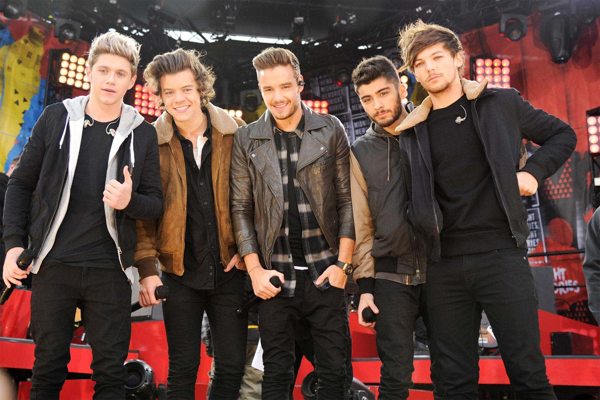 One Direction 桌布 壁紙 — 五位年輕男音樂家在燈光璀璨的演唱會舞台上自信地擺姿勢，身後有紅色燈光和設備。