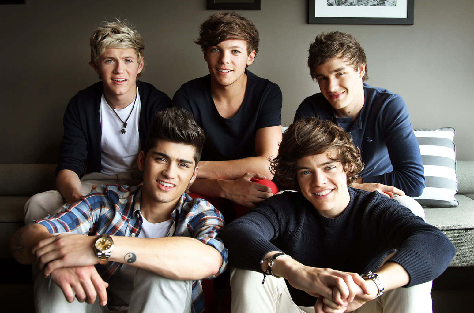 One Direction 桌布 壁紙 — 五位微笑的年輕男士在現代客廳的灰色沙發上隨意地一起擺姿勢。
