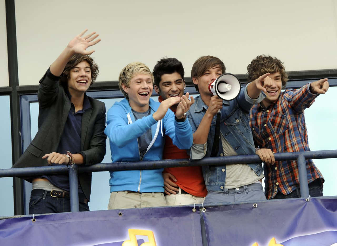 One Direction 桌布 壁紙 — 五位微笑的年輕男子穿著休閒服裝，在陽台欄杆上揮手並用擴音器歡呼。