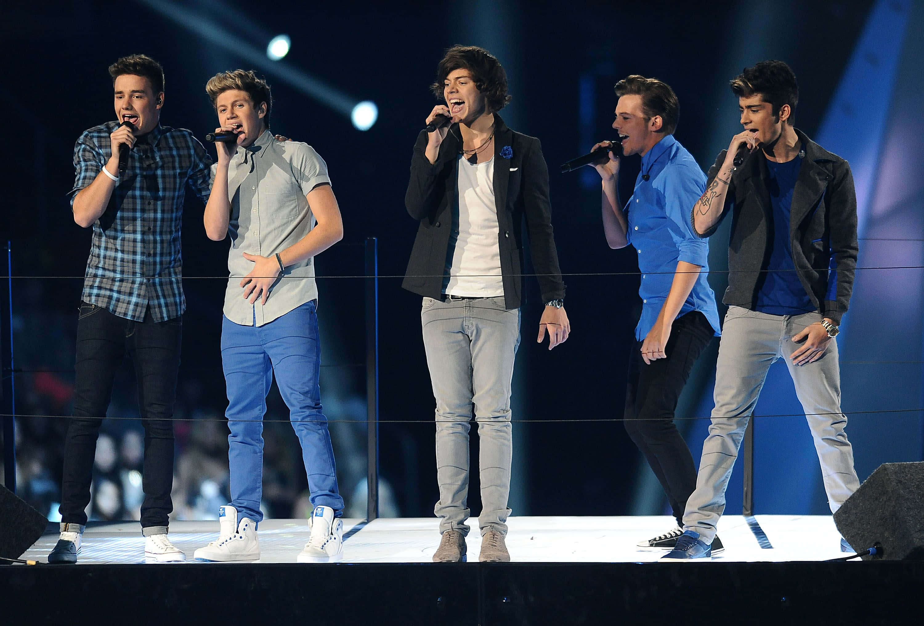 One Direction 桌布 壁紙 — 五位男歌手在燈光明亮的舞台上，在聚光燈下手持麥克風充滿活力地表演。