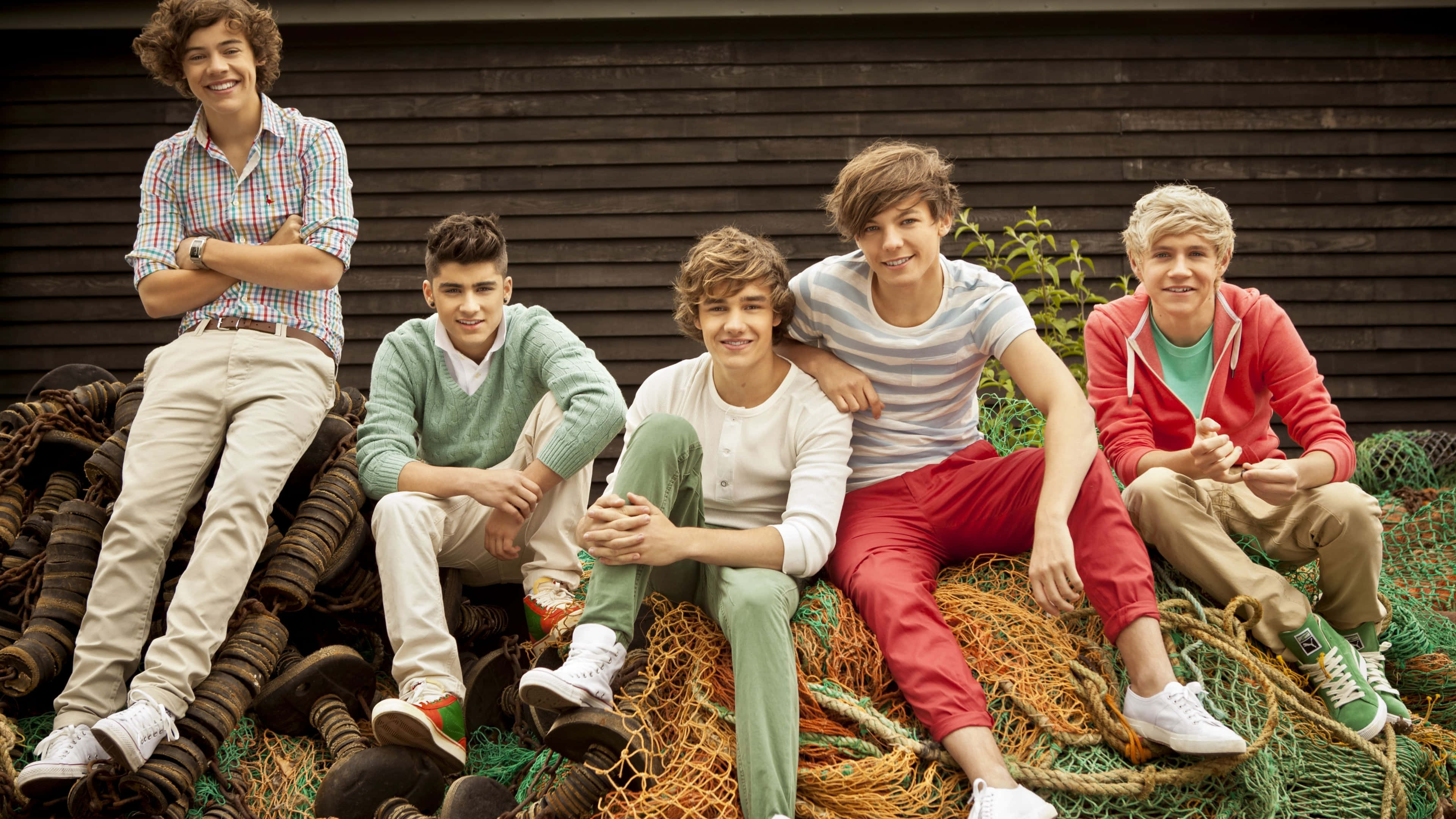 One Direction 桌布 壁紙 — 五位穿著休閒服裝的年輕男子微笑著，放鬆地坐在纏繞的漁網上，背景是深色木牆。