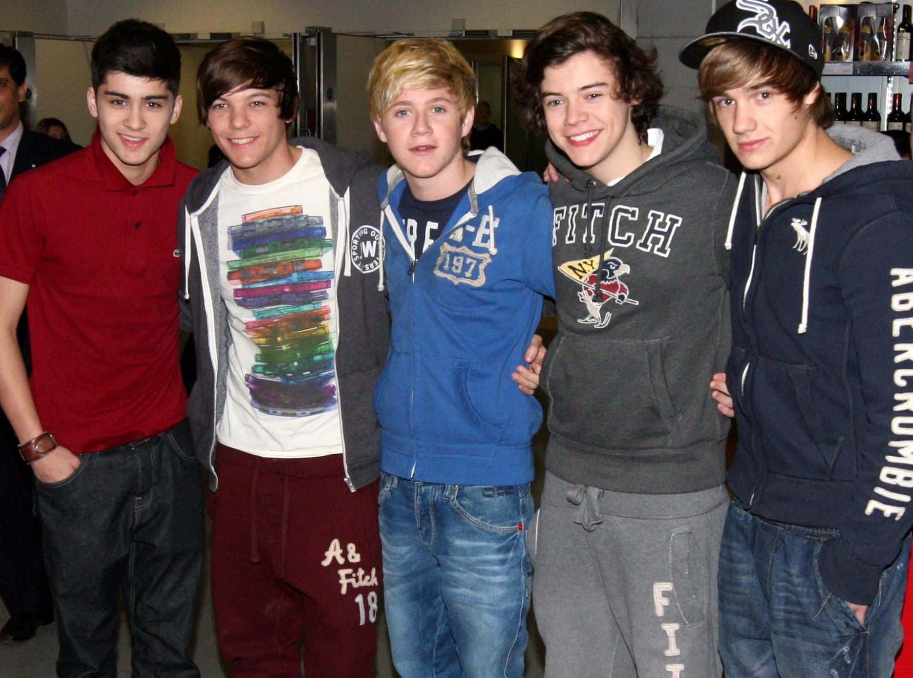 One Direction 桌布 壁紙 — 五位穿著休閒街頭服的年輕男子在溫暖燈光的室內環境中微笑合照。