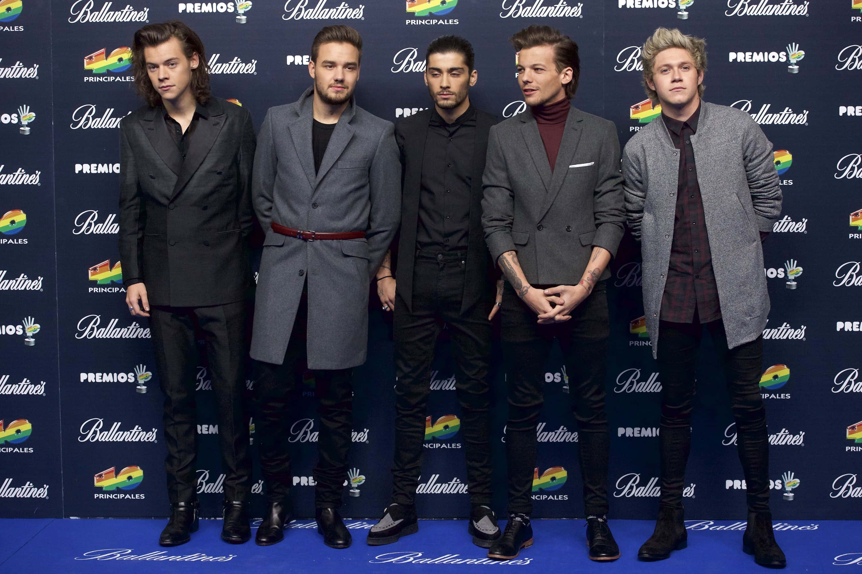 One Direction 桌布 壁紙 — 五位穿著訂製大衣的時尚男士在頒獎典禮上，自信地站在藍色地毯上，背景是品牌標誌。