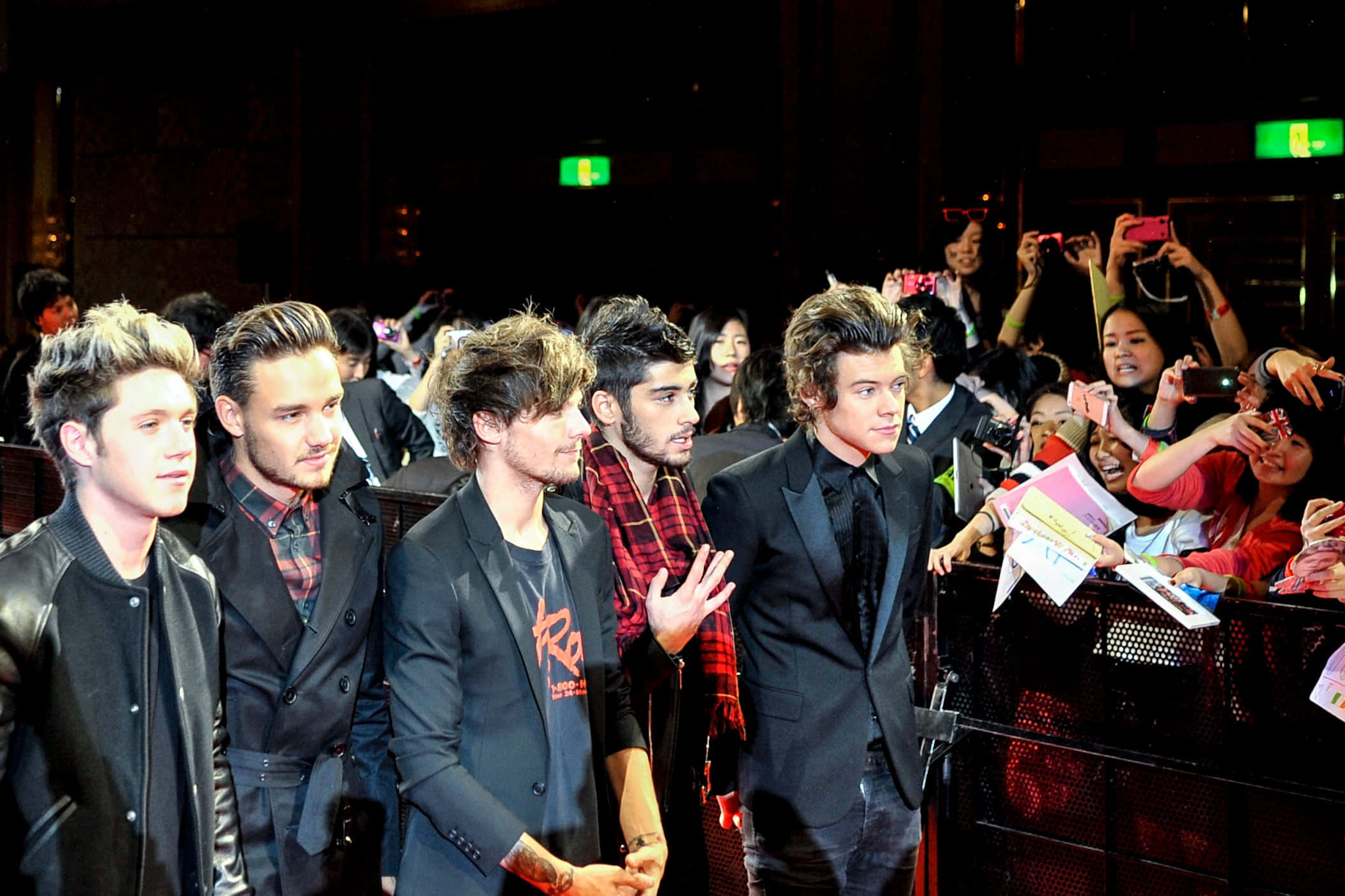 One Direction 桌布 壁紙 — 五位身穿時尚服裝的年輕男性在明亮的燈光下，於擁擠的室內活動中在圍欄後為粉絲擺姿勢。