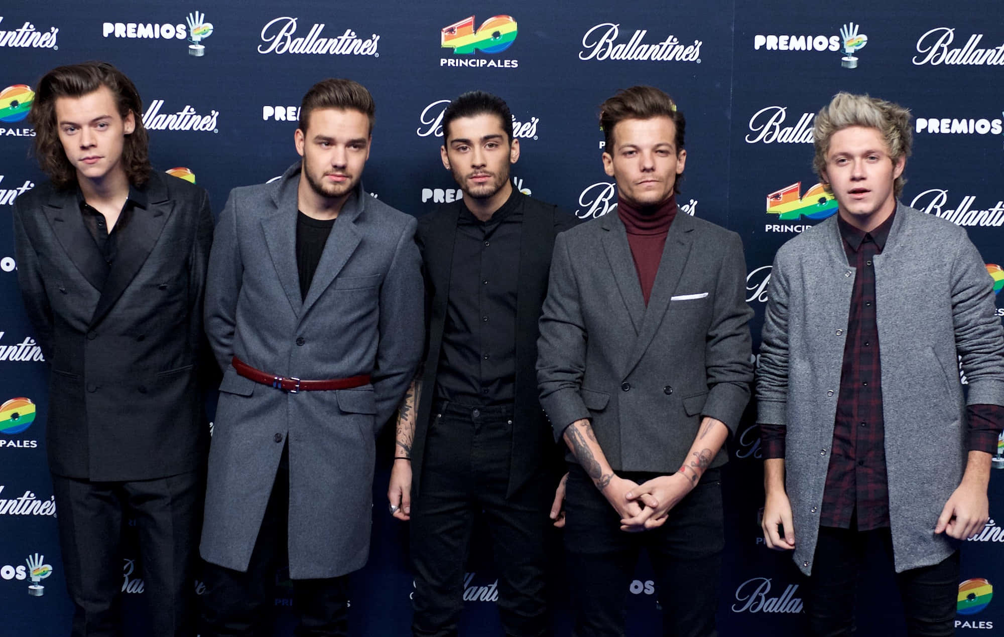One Direction 桌布 壁紙 — 五位身穿時尚西裝的男士在帶有標誌和彩虹色調的品牌活動背景前自信地擺姿勢。