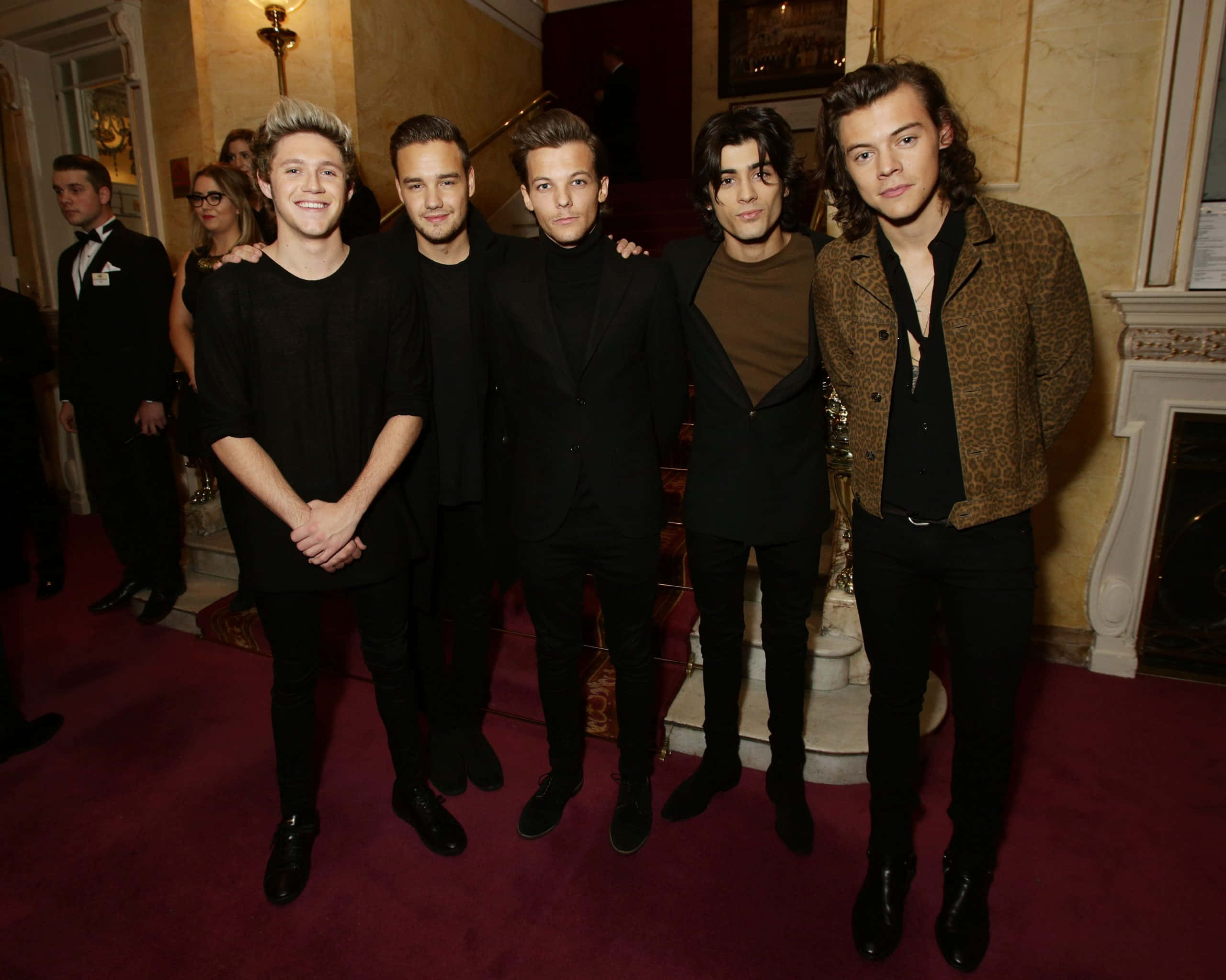 One Direction 桌布 壁紙 — 五位身穿時尚黑色服裝的年輕男子在優雅的室內環境中一起在紅地毯上擺姿勢。