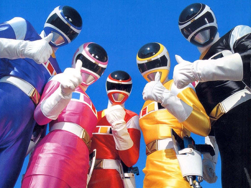 Power Rangers 桌布 壁紙 — 五位身穿藍色、粉紅色、紅色、黃色和黑色戰衣的恐龍戰隊在晴朗的藍天下自信地擺姿勢。