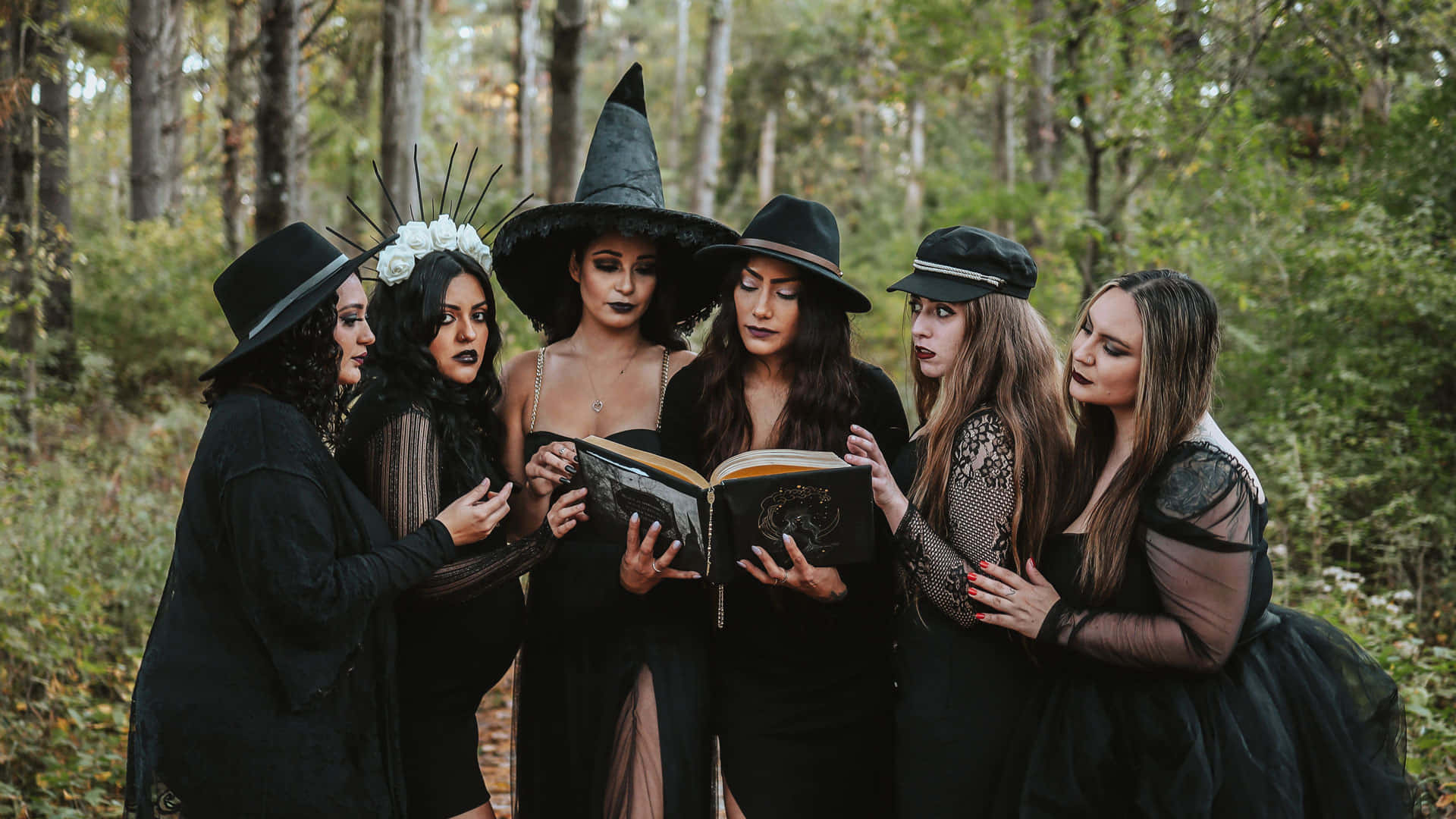 Grimoire 桌布 壁紙 — 五位身穿黑色女巫服裝的女性聚集在森林中，專注地研究一本打開的咒語書。