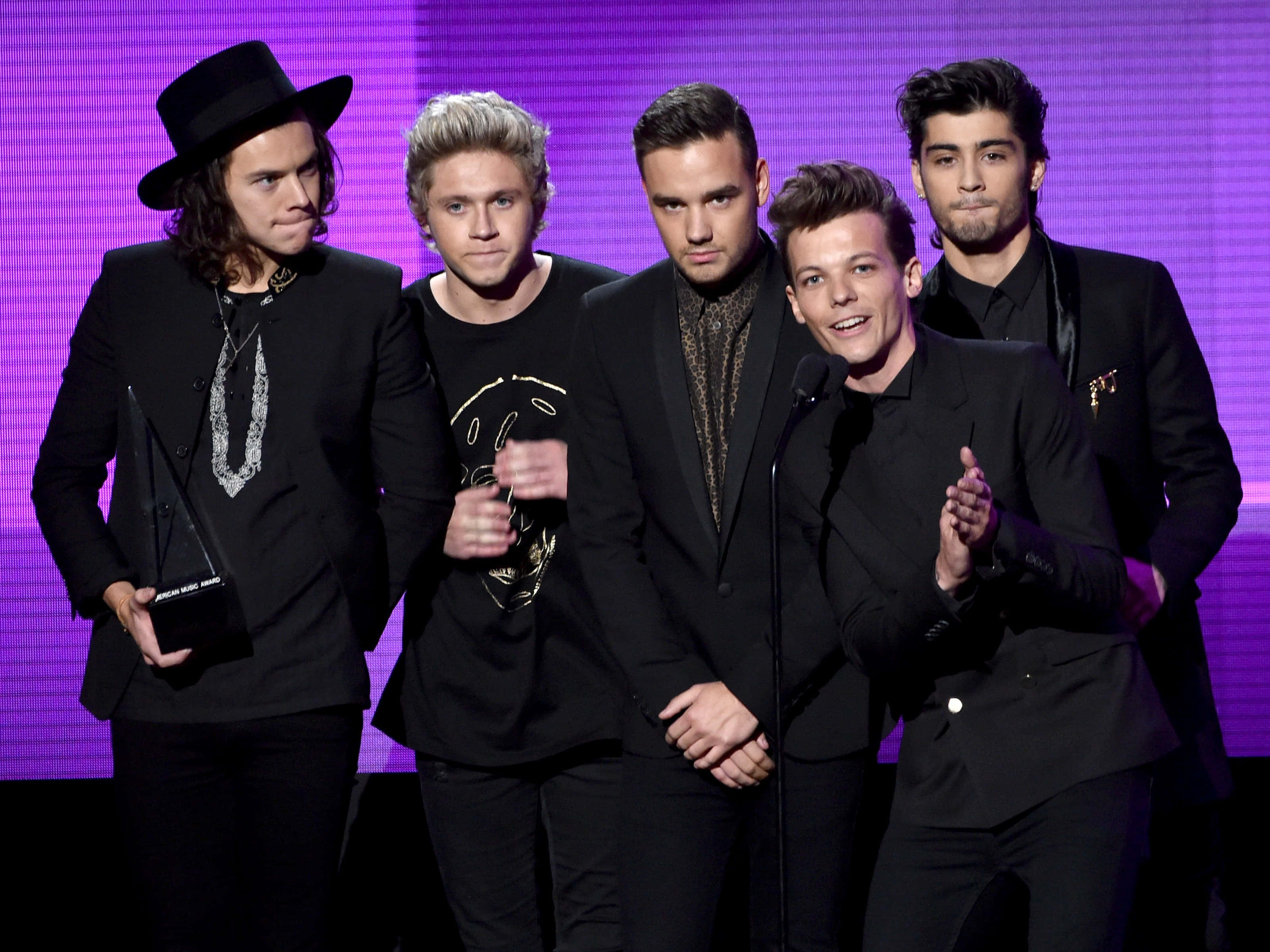 One Direction 桌布 壁紙 — 五位身穿黑色西裝的男子站在紫色燈光的舞台上，手持獎項並在頒獎典禮上鼓掌。