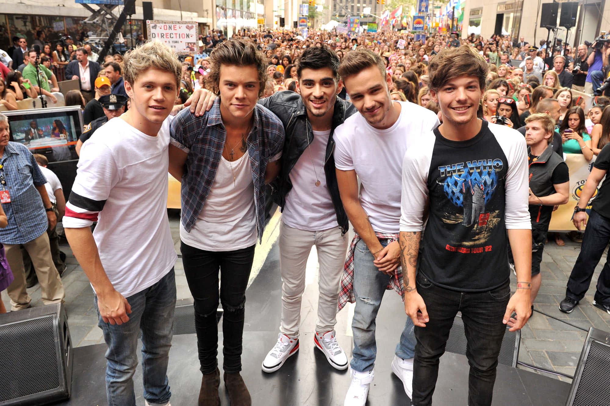 One Direction 桌布 壁紙 — 五位面帶微笑的年輕男士穿著休閒街頭服飾，在繁忙的城市活動中一起擺姿勢，面前是歡呼的人群。