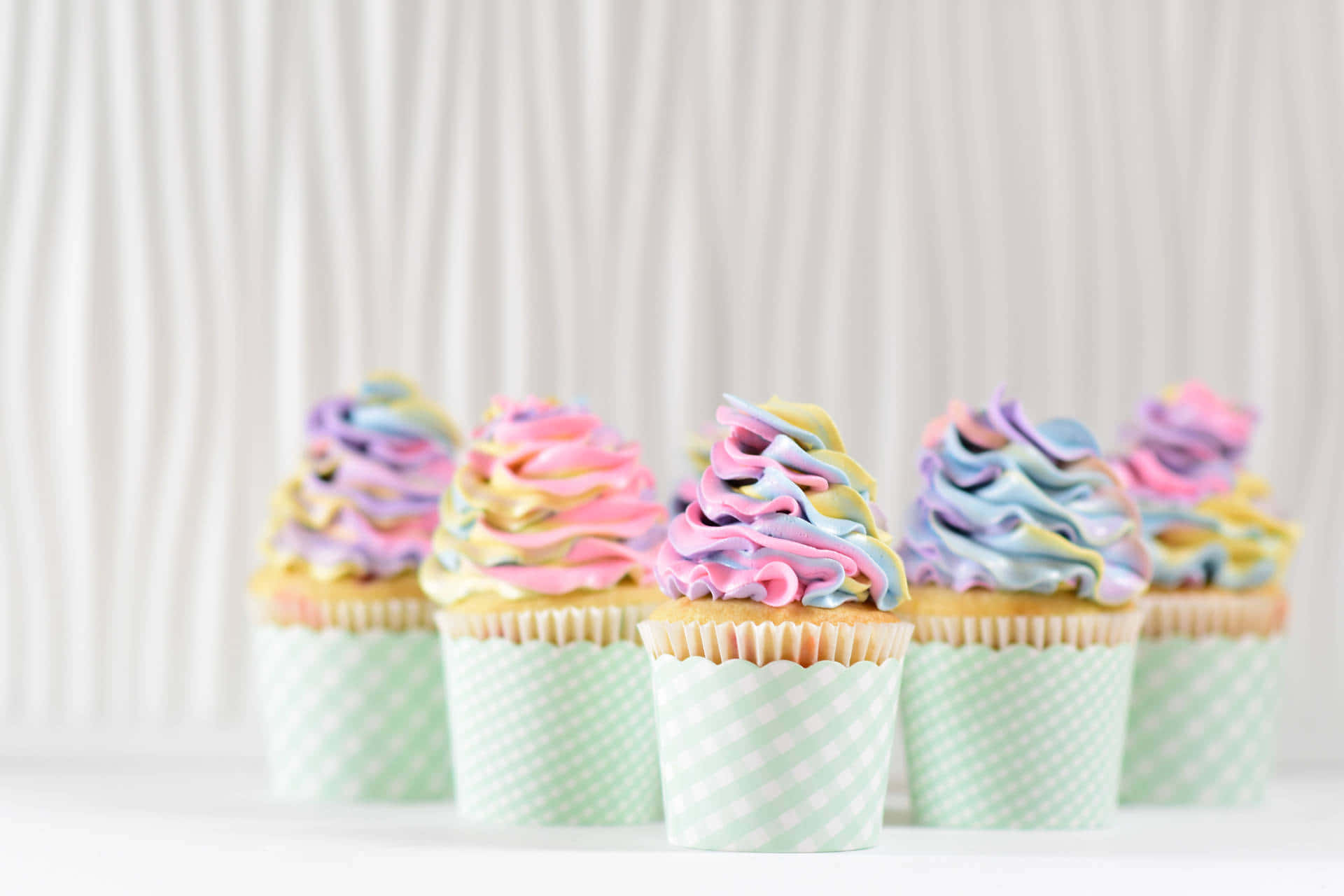 Cute Cupcake 桌布 壁紙 — 五個帶有柔和彩虹糖霜的紙杯蛋糕，放在格子襯紙中，置於白色表面上，背景為柔軟條紋。