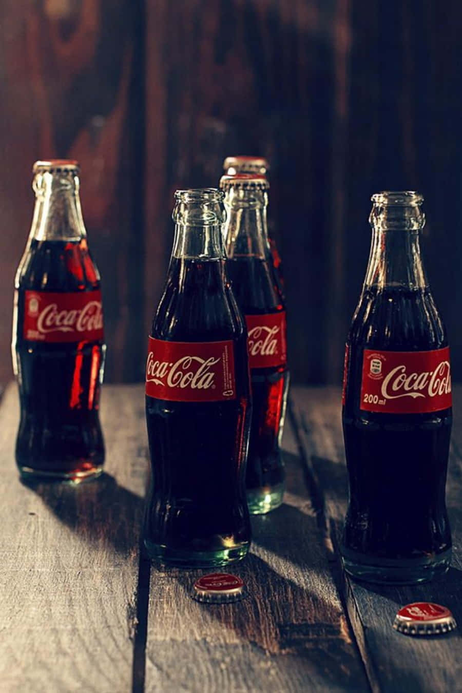 coca cola 桌布 壁紙 — 五個帶有紅色標籤的復古可口可樂玻璃瓶放在一張風化的木桌上，附近散落著瓶蓋。