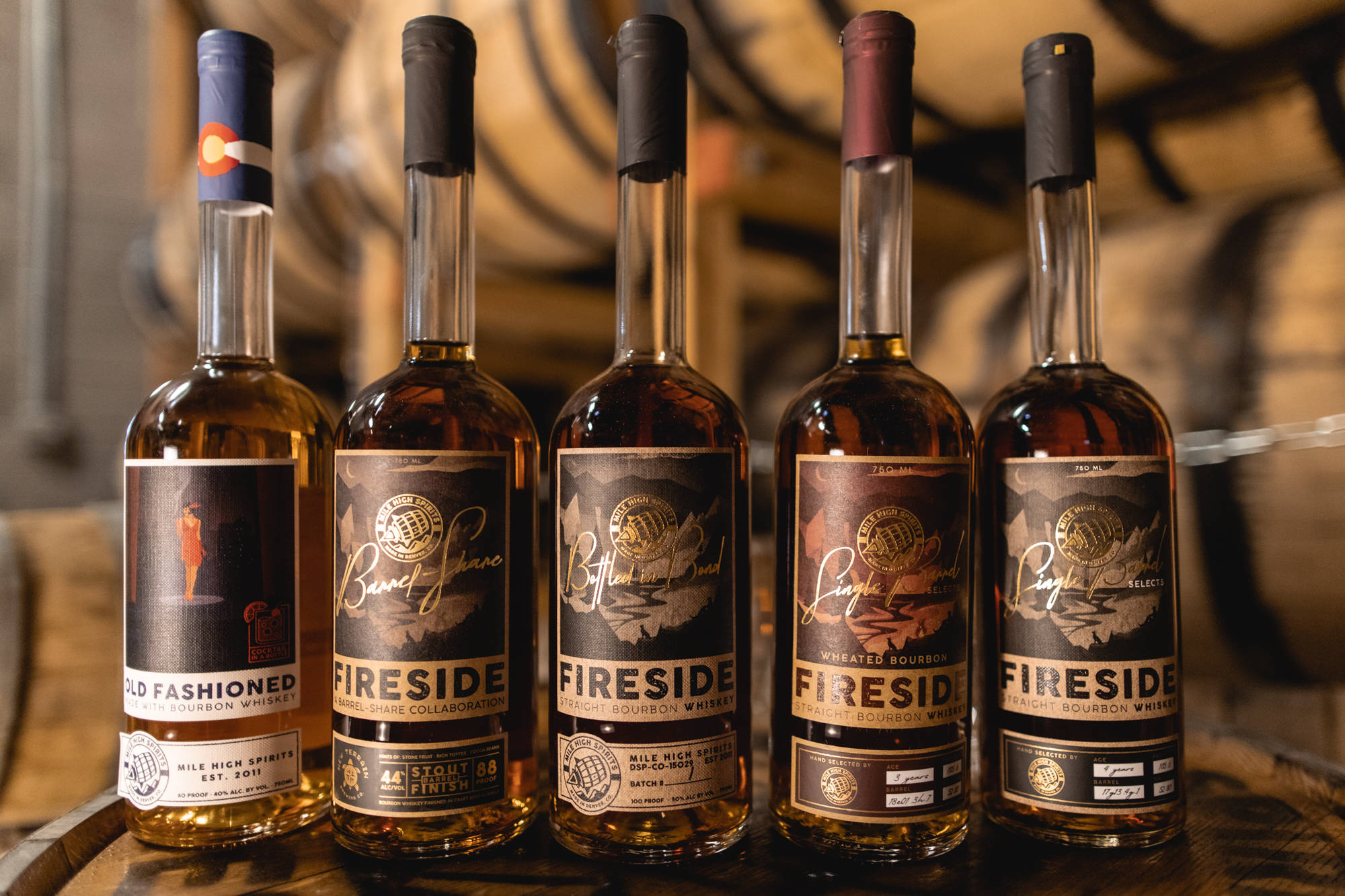 Mile High Spirits Different Fireside 壁紙 — 五個琥珀色波本威士忌酒瓶，帶有詳細標籤，排列在一個鄉村風格釀酒廠的木桶上。