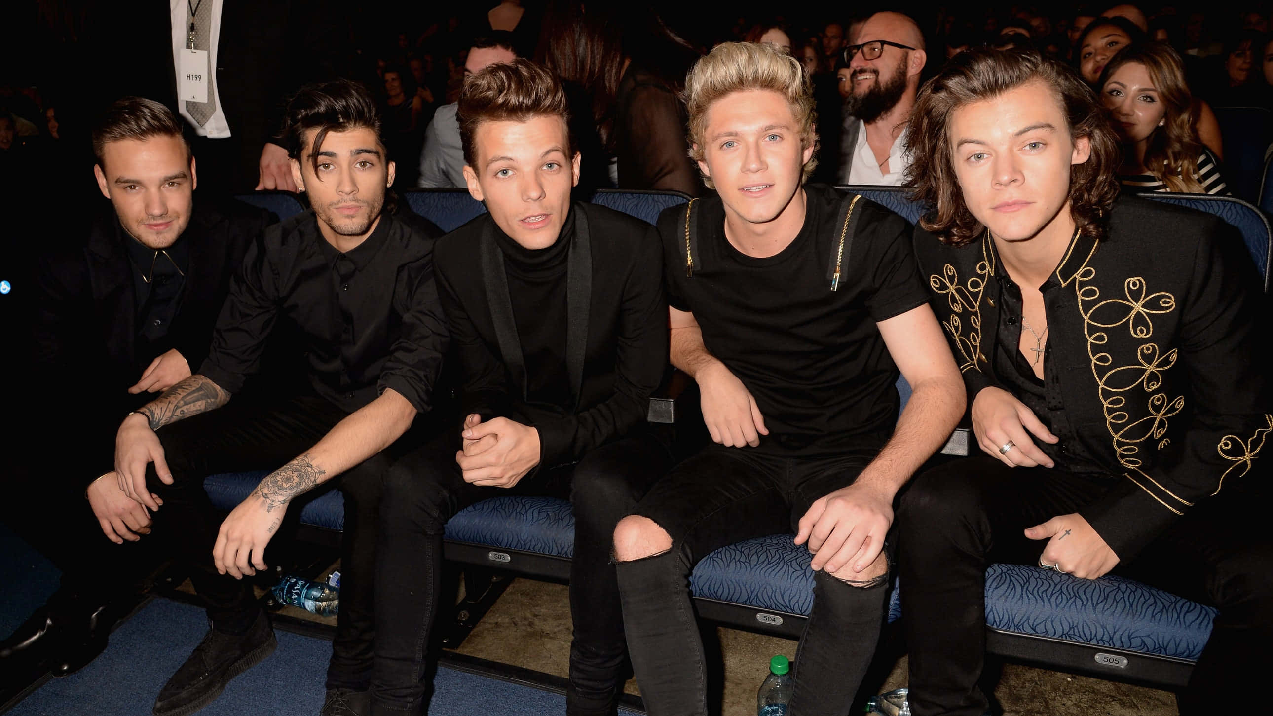 One Direction 桌布 壁紙 — 五個穿著黑色服裝的男人一起坐在藍色體育場座位上，參加一個燈光昏暗的活動，背後有群眾。