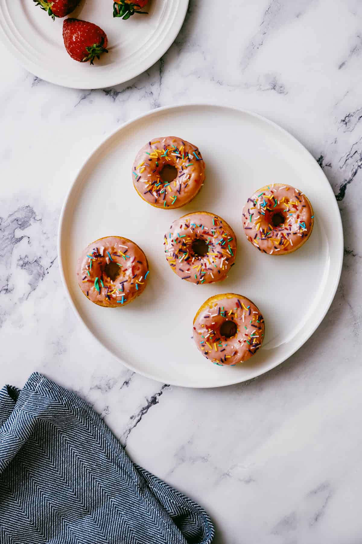Glazed Donut 桌布 壁紙 — 五個粉紅色糖霜甜甜圈，上面有彩色糖粒，放在白色盤子上，置於大理石檯面，旁邊有草莓和藍色布。