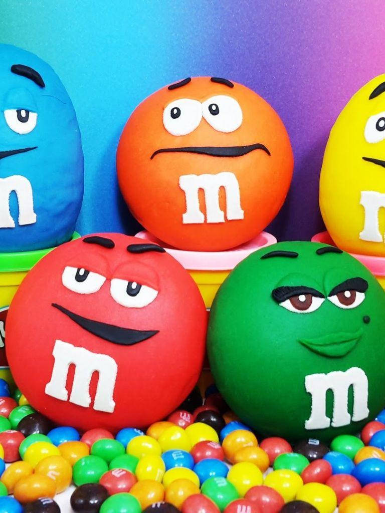 Chocolate 桌布 壁紙 — 五個色彩繽紛的M&M's角色，表情豐富，坐在各種糖果上，背景是充滿活力的漸變色。