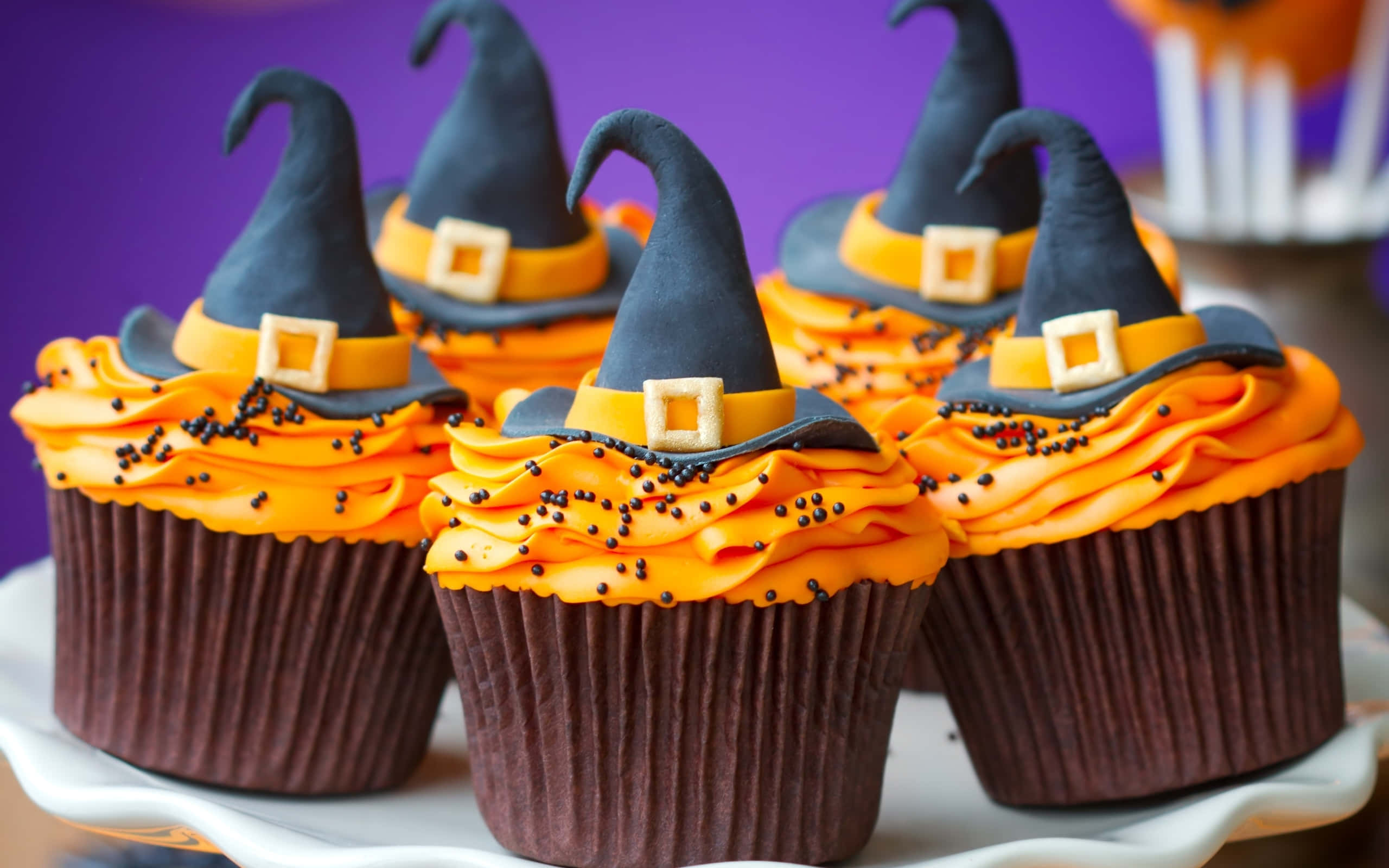 Halloween Cupcakes 桌布 壁紙 — 五個萬聖節紙杯蛋糕，頂部有巫婆帽和橙色糖霜，放在白色碟子上，背景為紫色。