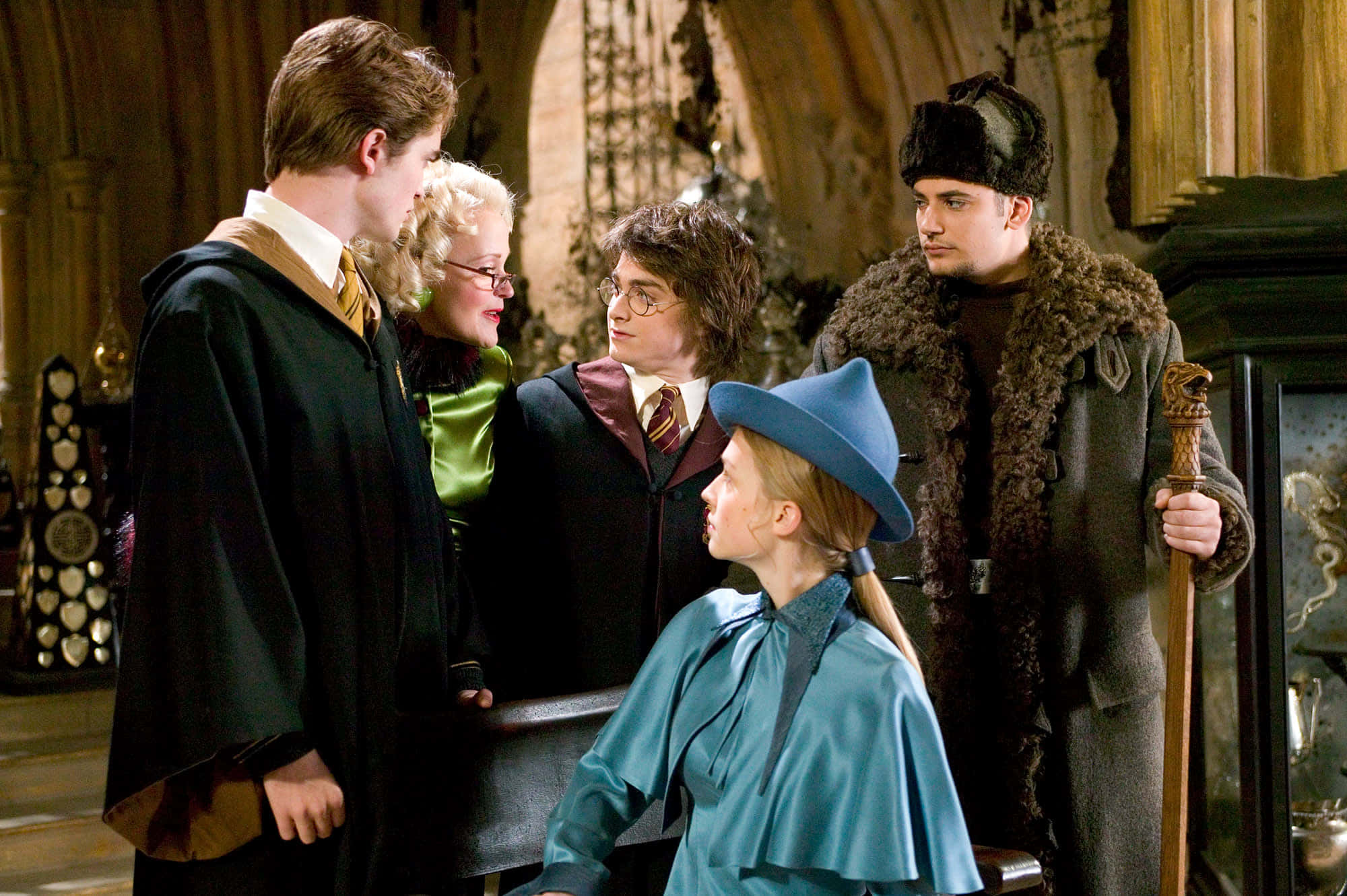 The Triwizard Cup 桌布 壁紙 — 五個身穿巫師袍的角色在一個帶有拱形窗戶和華麗裝飾的宏偉石廳中交談。
