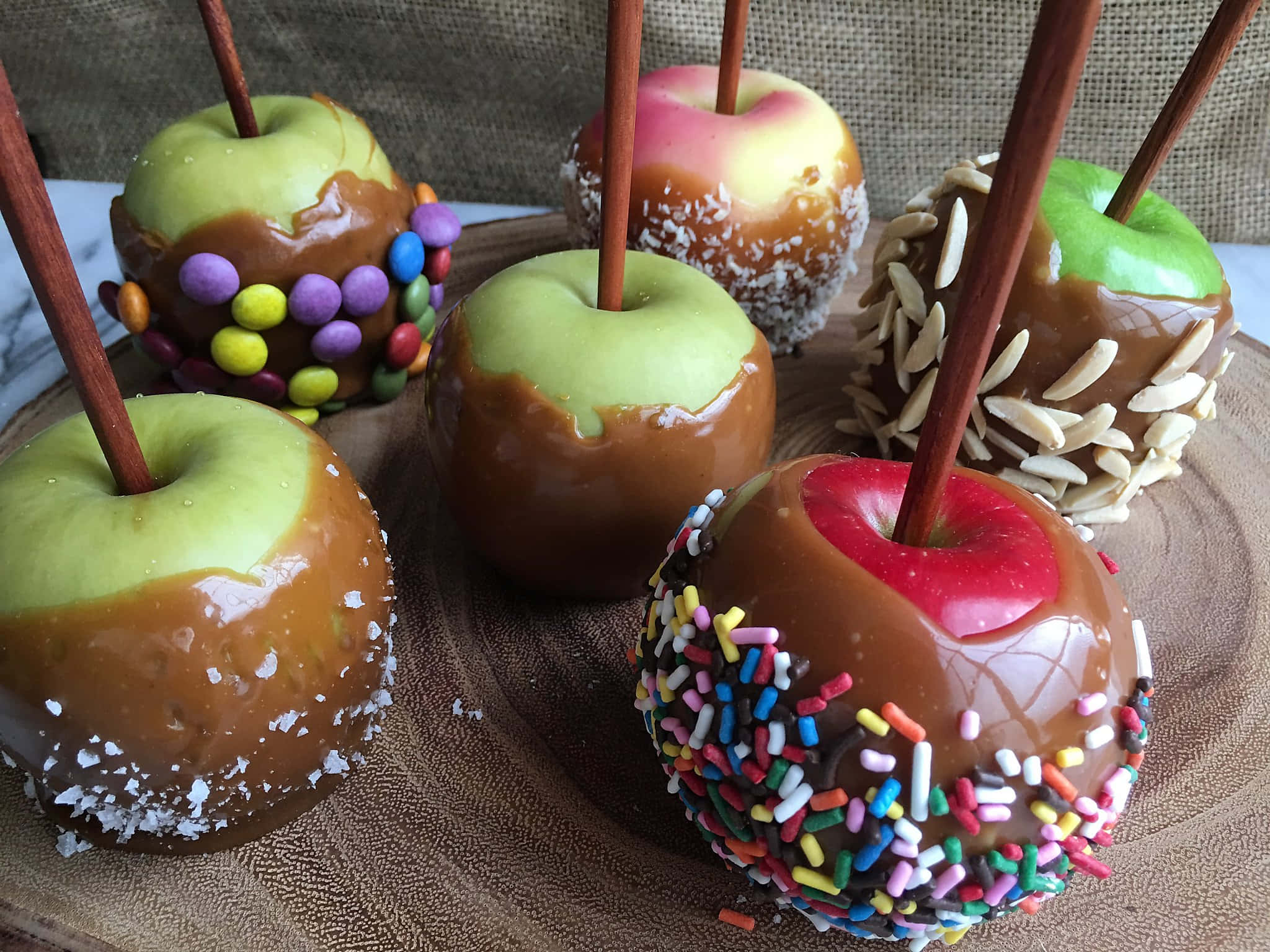 Caramel Apples 桌布 壁紙 — 五個閃亮的焦糖蘋果，上面有彩色配料如糖粒和杏仁，放在木板上。