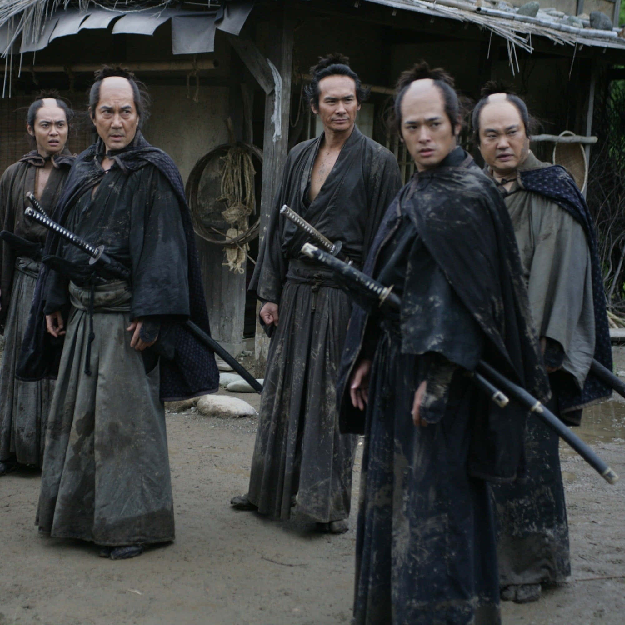 Samurai Movies 桌布 壁紙 — 五個面容陰沉的武士穿著破爛的深色長袍，站在泥濘的庭院中，面前是一座茅草屋頂的質樸木建築。