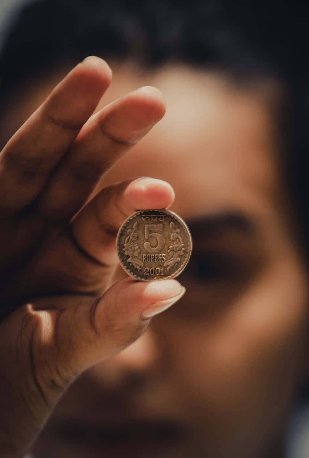 五分硬幣 壁紙 — 一隻手拿著一枚2001年的5盧比印度硬幣，對焦清晰，背景是一張柔和模糊的人臉。