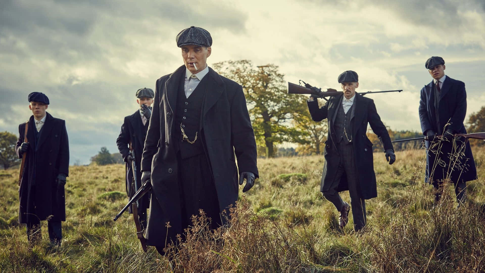 Peaky Blinders 桌布 壁紙 — 五名戴著復古平頂帽、身穿乾濕褸的男子，在陰天下手持步槍走過草地。