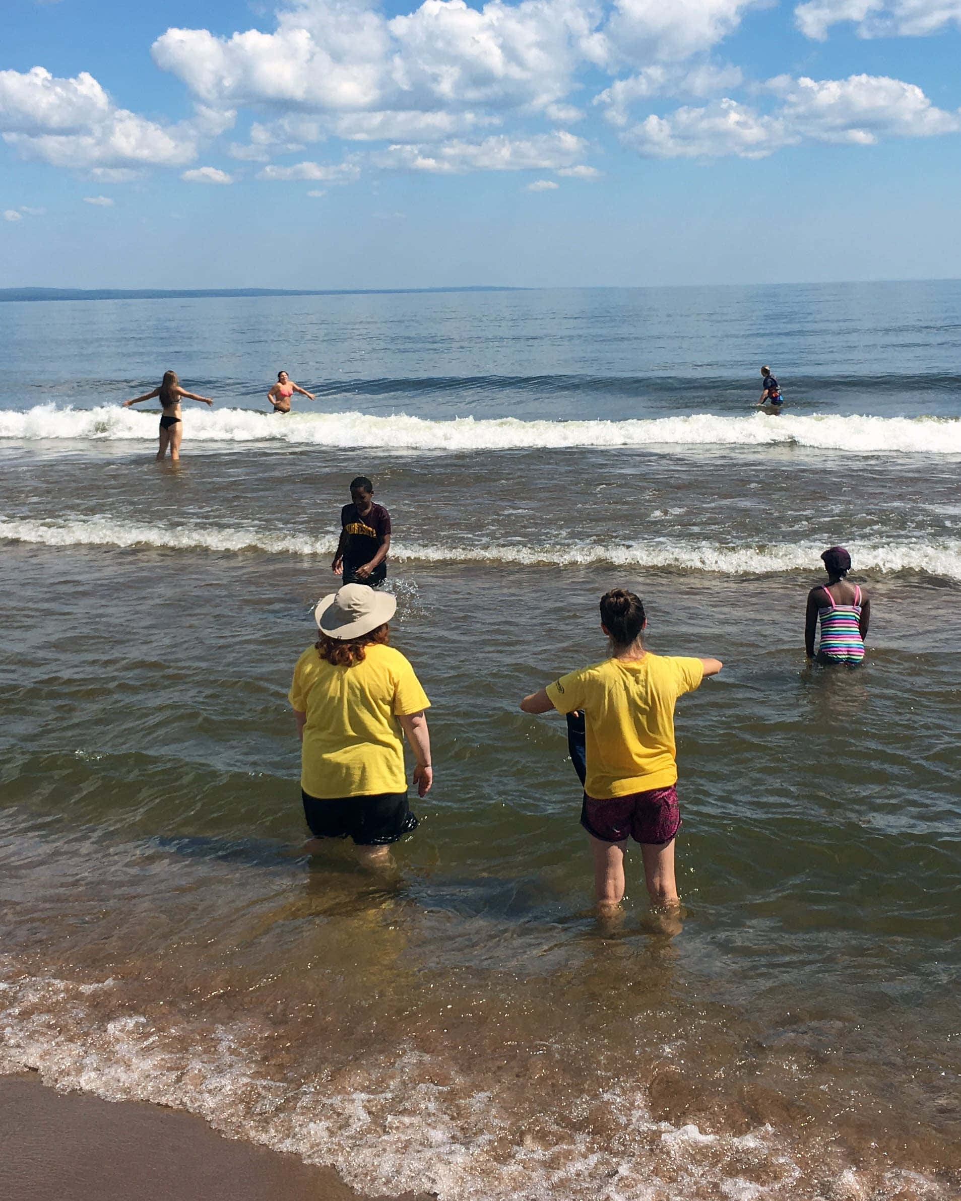 五大湖遊客圖 壁紙 — 穿著黃色襯衫的人們站在淺海水中，而其他人在明亮的藍天白雲下衝浪溫和的海浪。