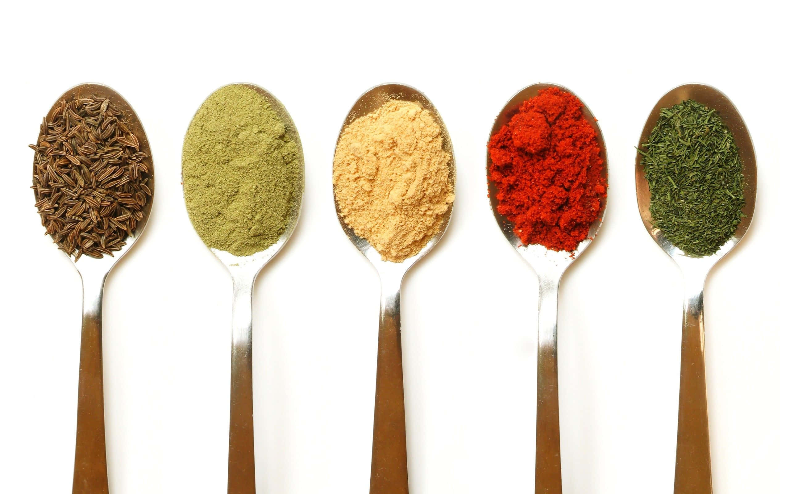 Spice 桌布 壁紙 — 五隻匙羹盛載著鮮豔的香料——棕色種子、綠色粉末、黃色粉末、紅辣椒碎和綠色香草——在純白色背景上。