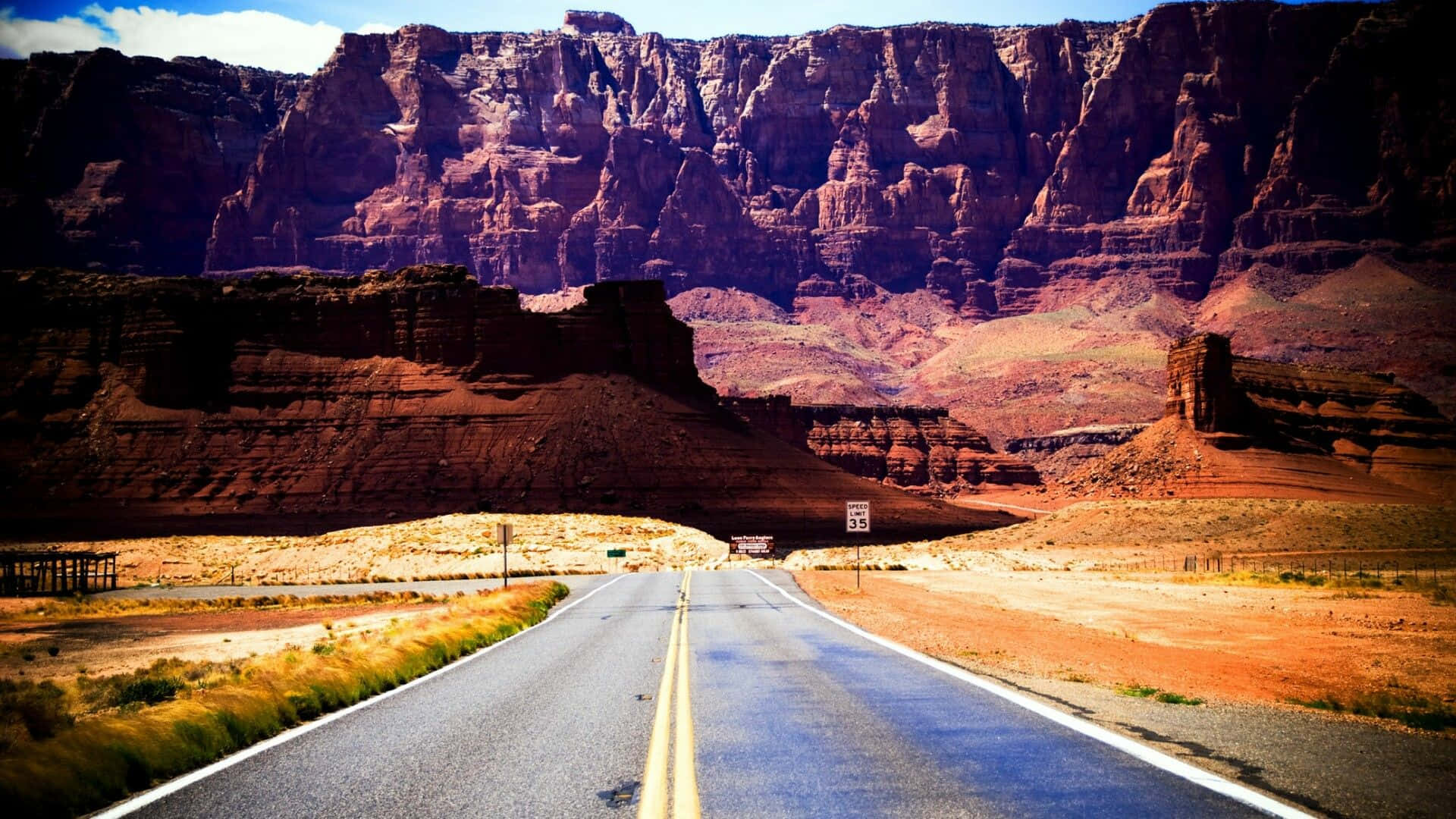 亞利桑那沙漠日落的壯麗景色 壁紙 — 一條筆直的瀝青道路在明亮的藍天下延伸向高聳的紅色岩壁。