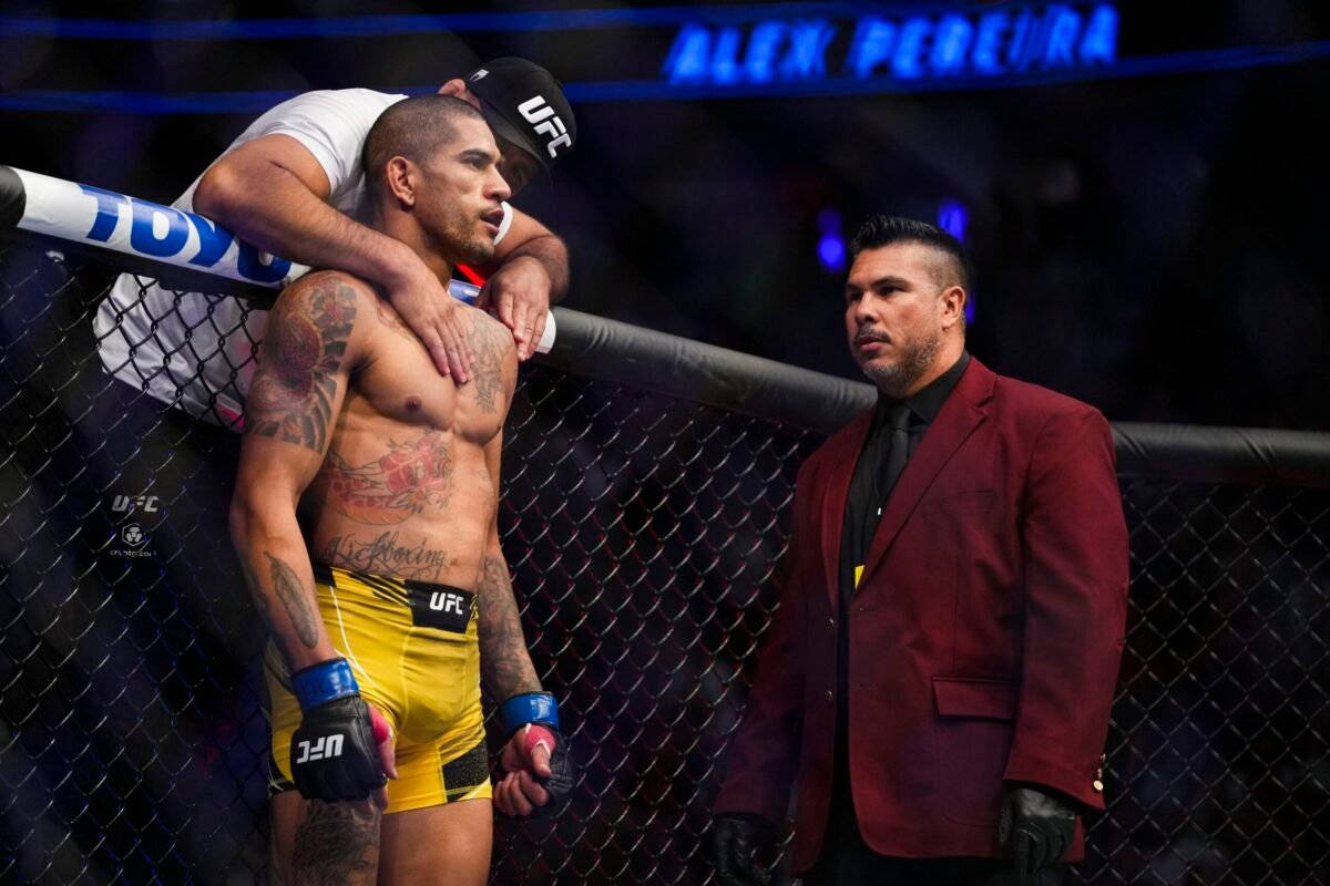 亞歷克斯·佩雷拉與教練同框 壁紙 — 一位有紋身的UFC拳手穿著黃色短褲站在八角籠內，而一名身穿紅色西裝的男子正專注地觀看。