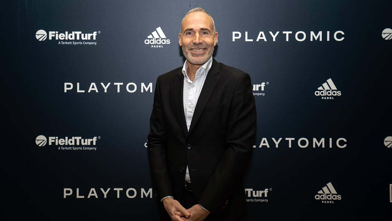 黑西裝男士在帶有 Playtomic FieldTurf 和 Adidas Padel 標誌的品牌活動背景前微笑。