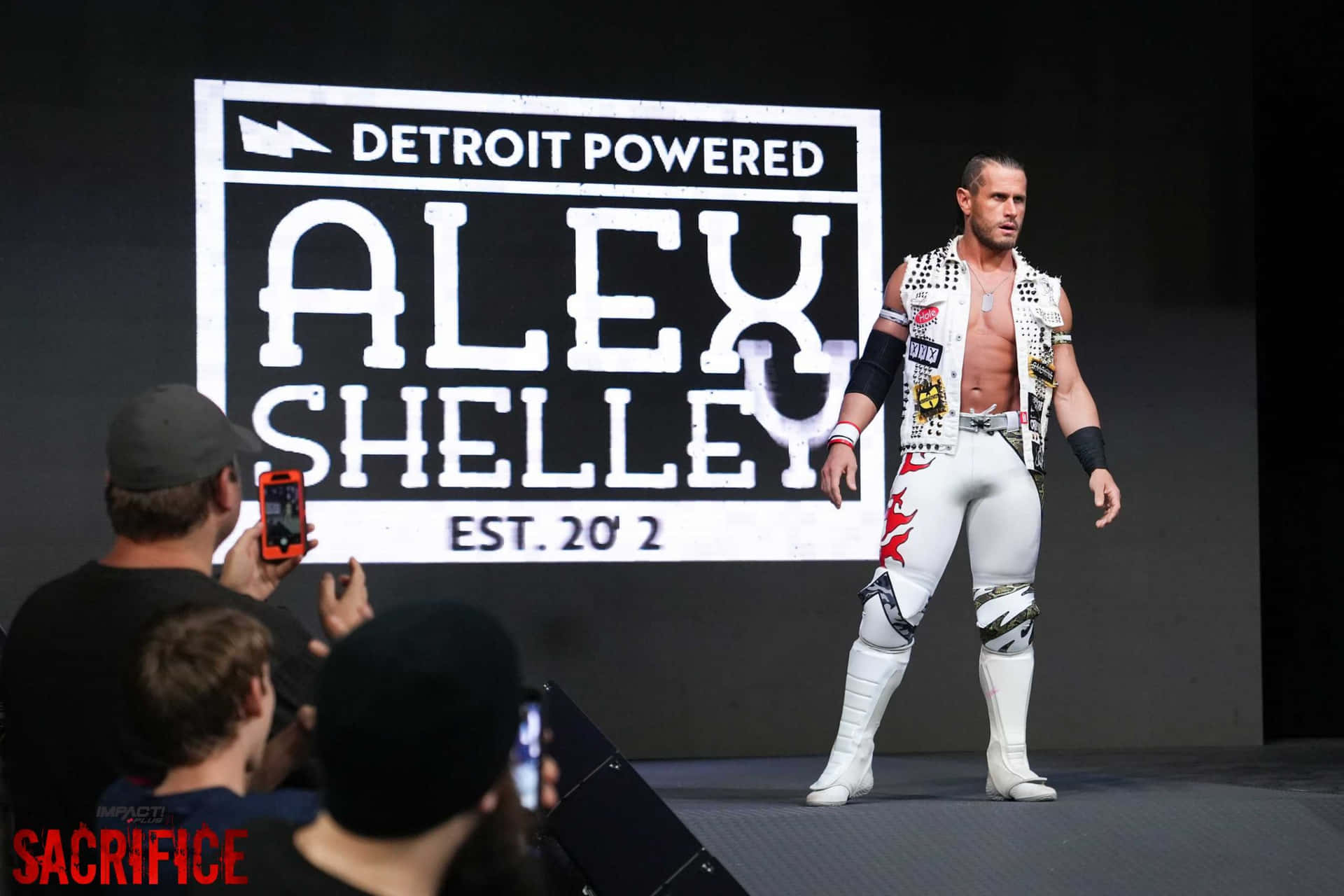 摔角手 Alex Shelley 身穿白色裝備，在 Impact Wrestling 活動中站在舞台上。