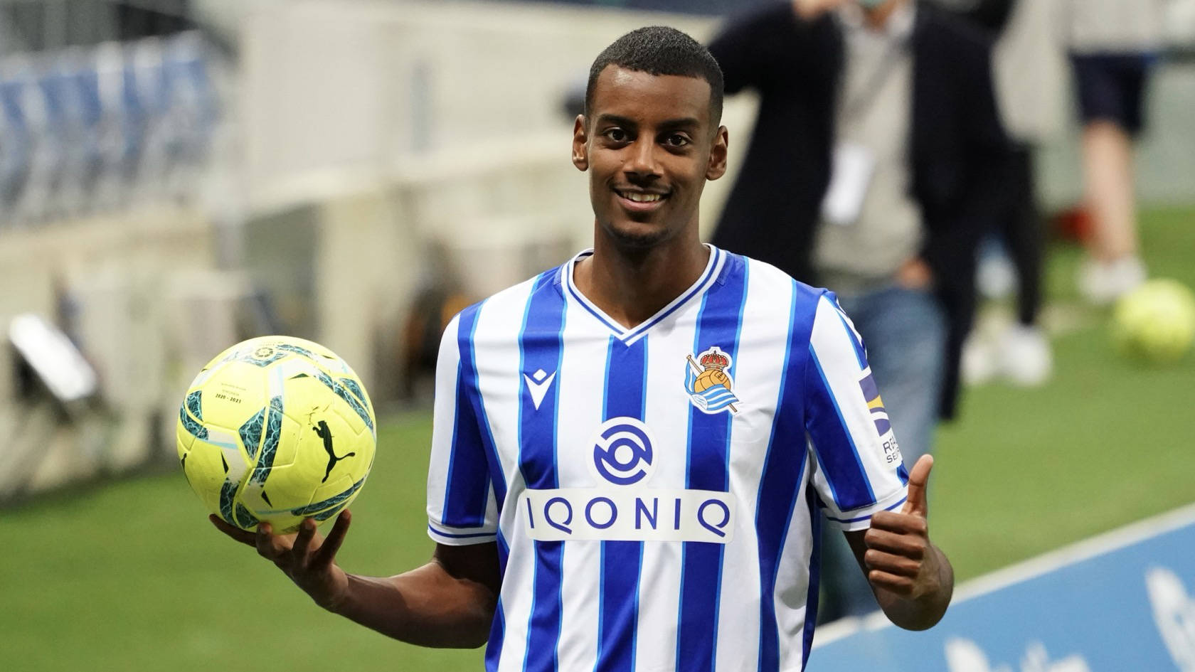 亞歷山大·伊薩克手持足球 壁紙 — 一位微笑的足球運動員身穿藍白條紋球衣，手持黃色足球，在綠色球場上豎起大拇指。