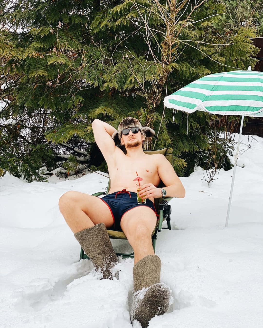 穿著泳褲和毛帽的男子在雪地上放鬆，有飲料和傘。