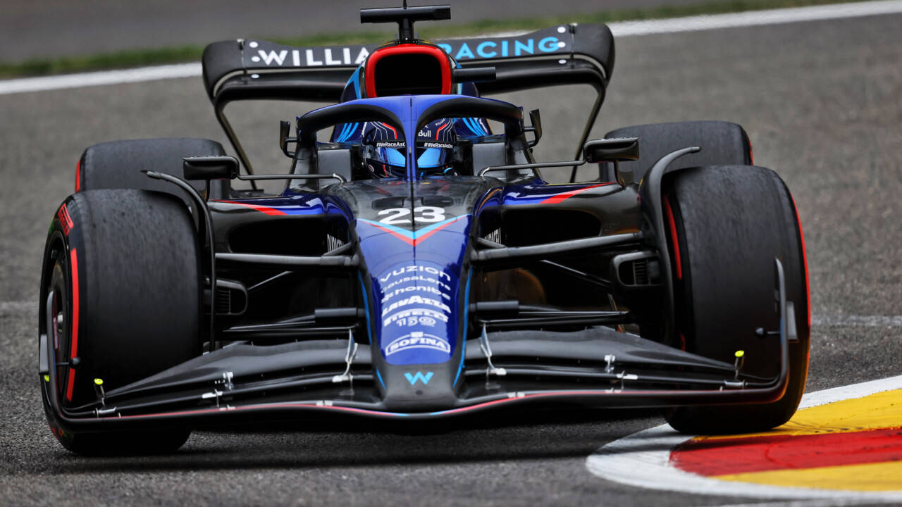亞歷山大·阿爾本駕駛藍色F1賽車 壁紙 — 一輛藍黑相間的F1賽車（編號23）在潮濕的賽道上行駛，靠近紅、黃、白色的路緣石。