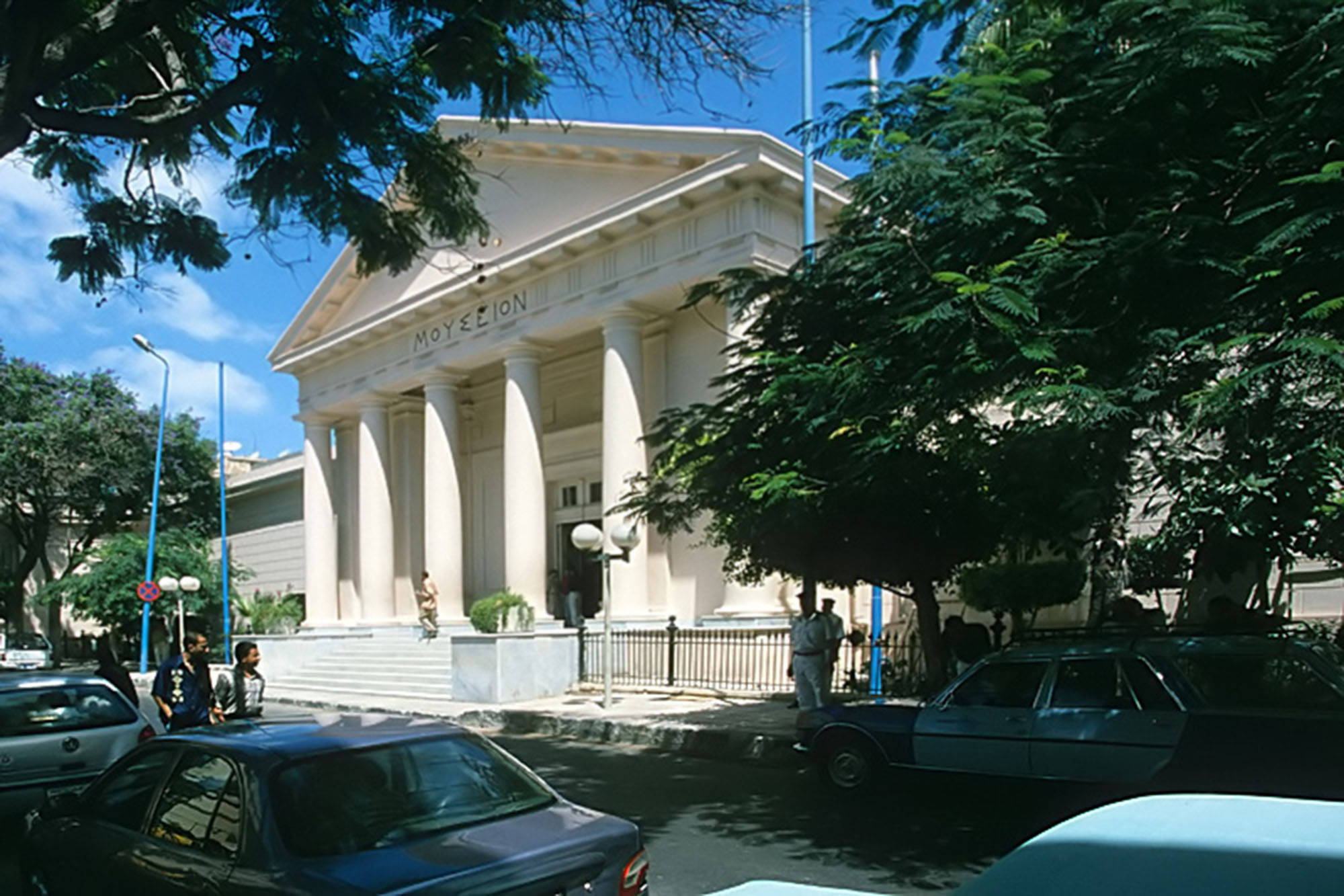 亞歷山大希臘羅馬博物館壯麗風景 壁紙 — 一座宏偉的新古典主義建築，擁有高大的柱子和三角形山牆，矗立在明亮的藍天下，周圍環繞著茂密的綠樹和停泊在陽光普照街道上的汽車。