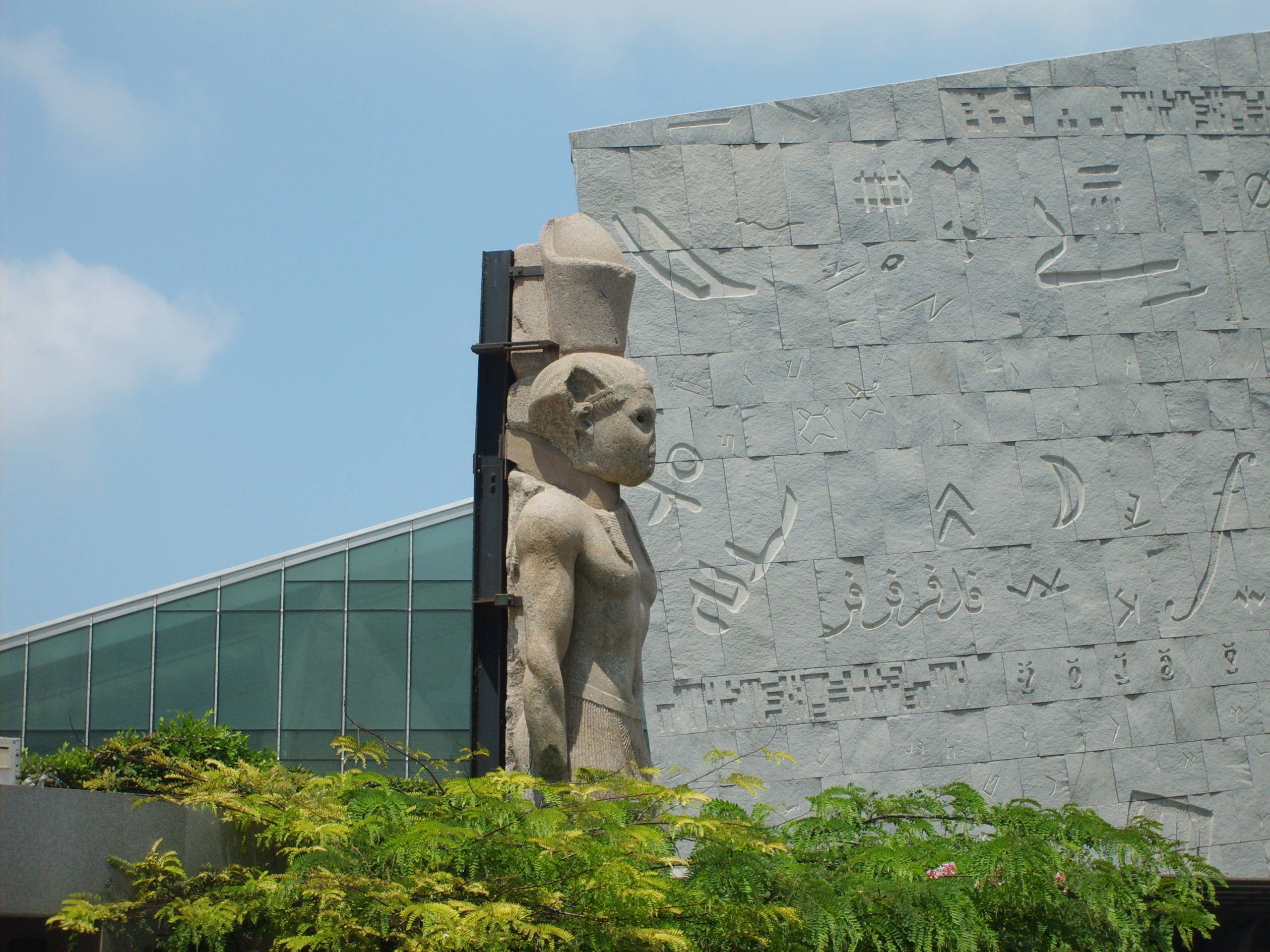 亞歷山大托勒密雕像 壁紙 — 古埃及雕像矗立在一座現代建築前，其石質外牆上刻有象形文字，背景是清澈的藍天。