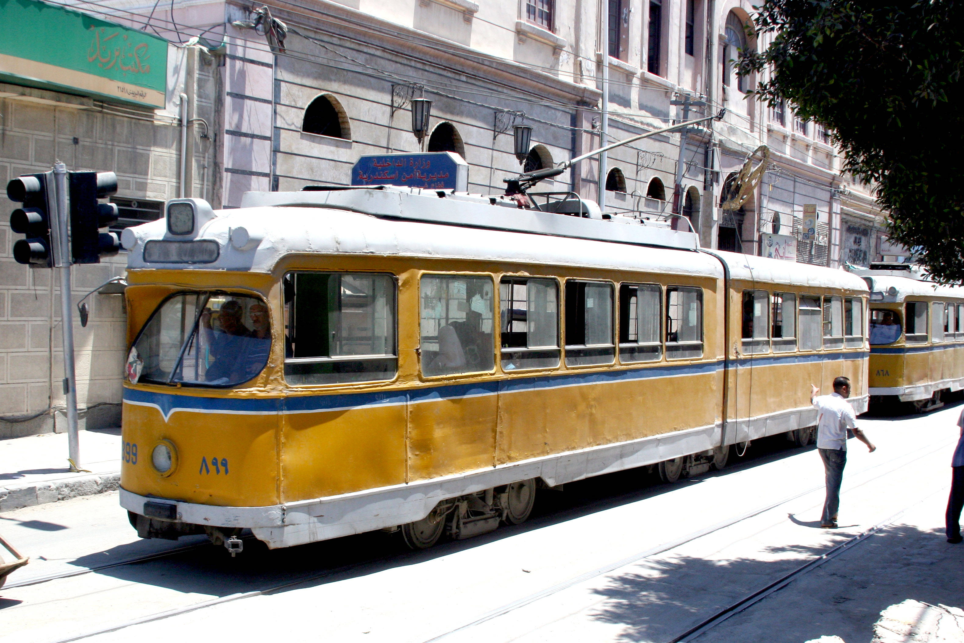 亞歷山大黃色電車 壁紙 — 一輛帶有藍色飾邊的復古黃色電車在陽光明媚的城市街道上滑行，經過歷史建築和行人。