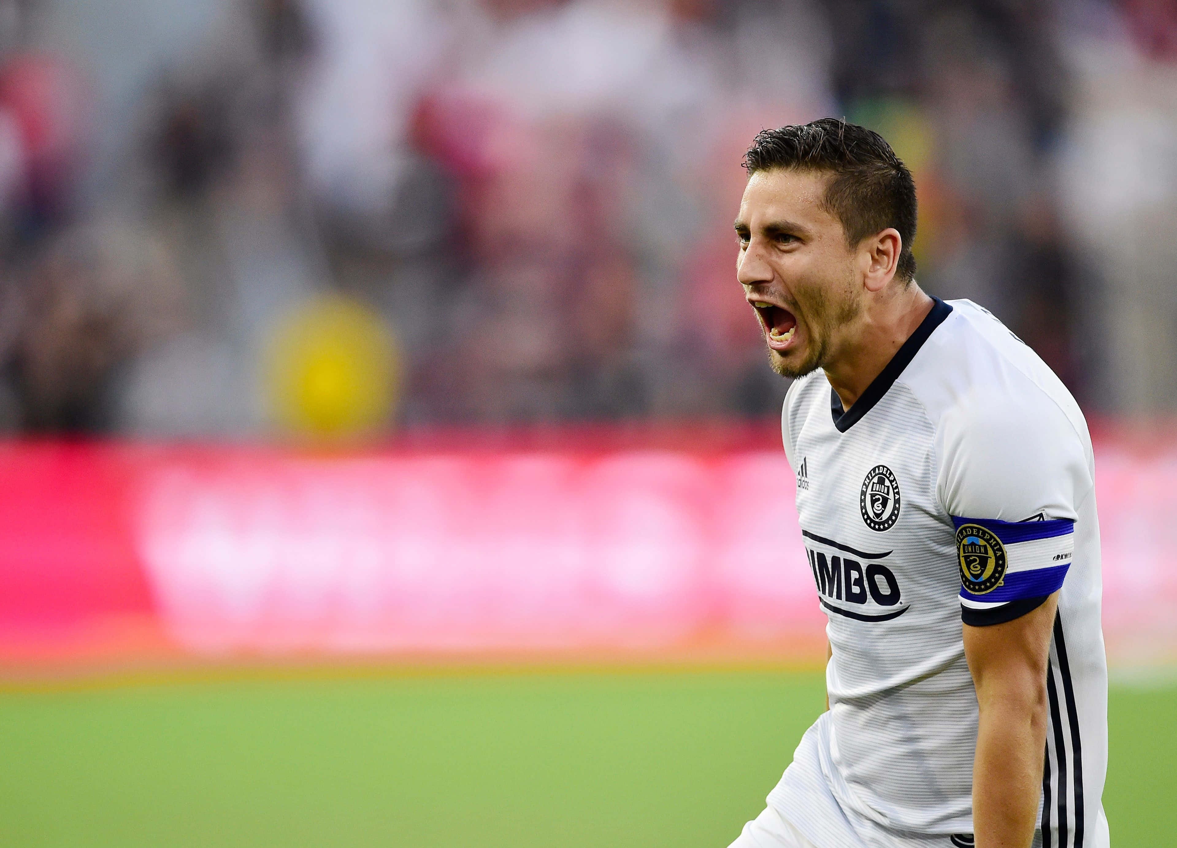 亞歷杭德羅·貝多亞對陣 D.C. United 壁紙 — 一名身穿白色球衣的足球運動員在充滿活力的綠色球場上勝利吶喊，身後是模糊的體育場人群。