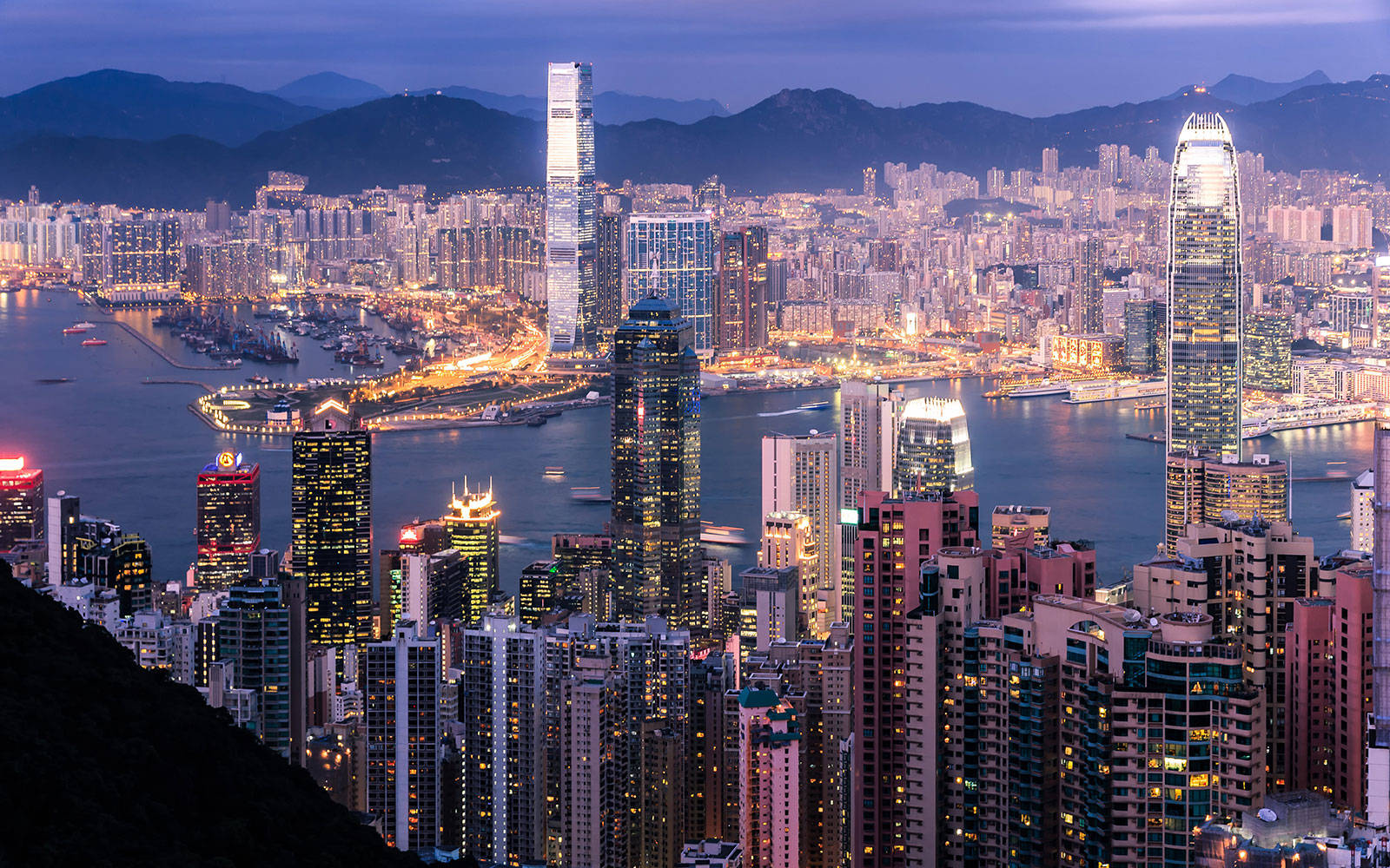 亞洲城市香港天際線 壁紙 — 香港天際線的黃昏全景，發光的摩天大樓和港口船隻映襯著黑暗的山脈。