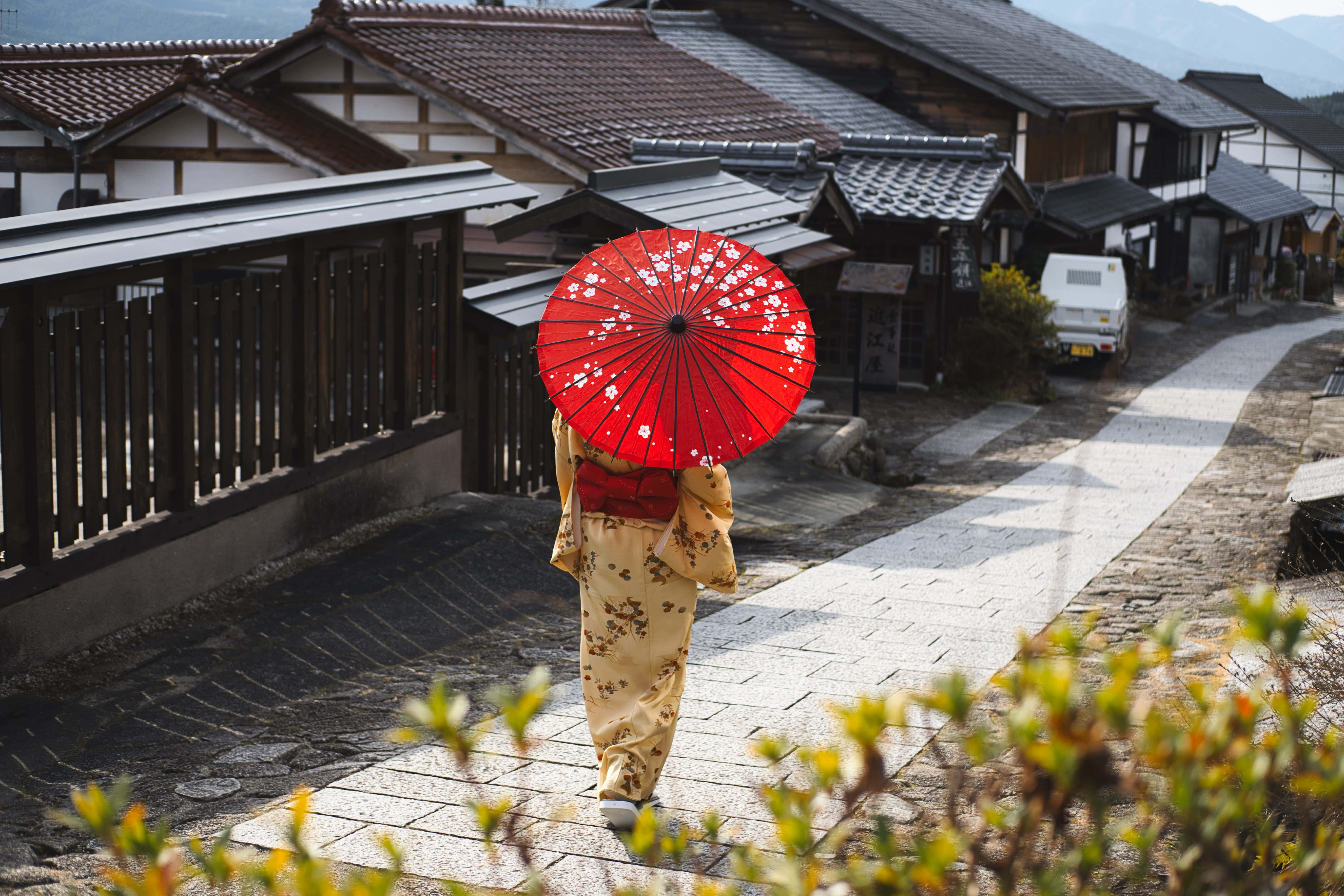 亞洲天際線之美 壁紙 — 在傳統日本村莊中，一位穿著花卉和服的人撐著紅色櫻花傘沿著石徑走遠。
