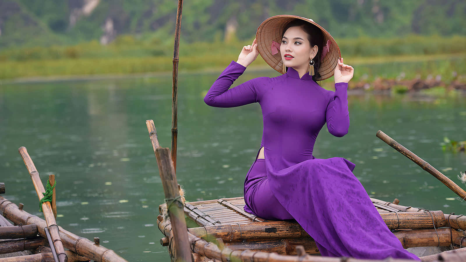 亞洲女性坐於木船 壁紙 — 一位身穿鮮豔紫色奧黛和錐形帽的女子優雅地坐在漂浮在寧靜綠色湖面上的竹筏上。