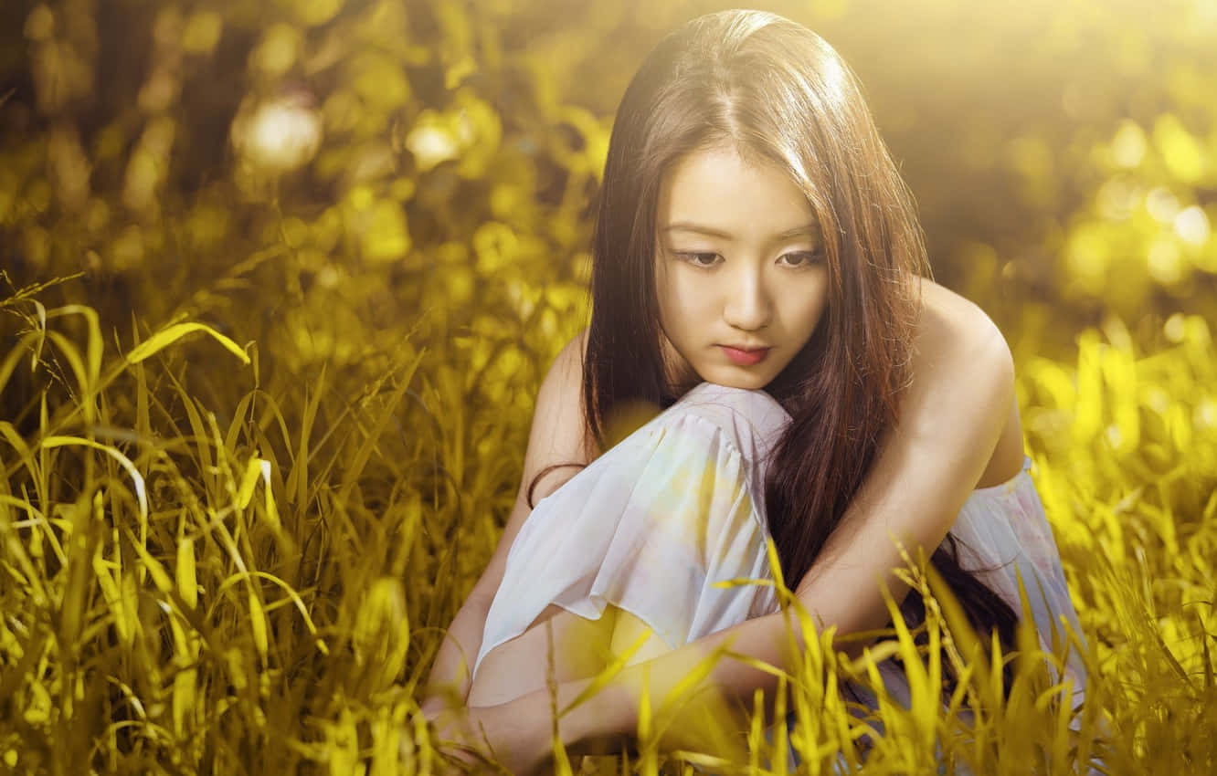 亞洲女性坐於草地 壁紙 — 一位年輕女性坐在金色草叢中，沐浴在溫暖陽光下，帶著沉思的表情。