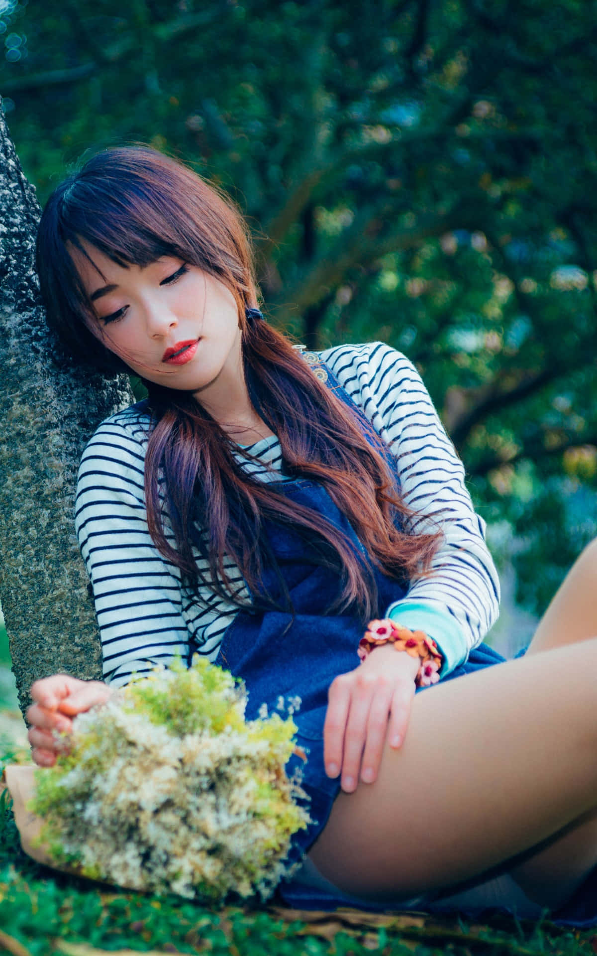 亞洲女性持花束 壁紙 — 一位長著棕色長髮的年輕女子靠著樹坐著，在茂密的綠色森林中手持一束苔蘚花束。