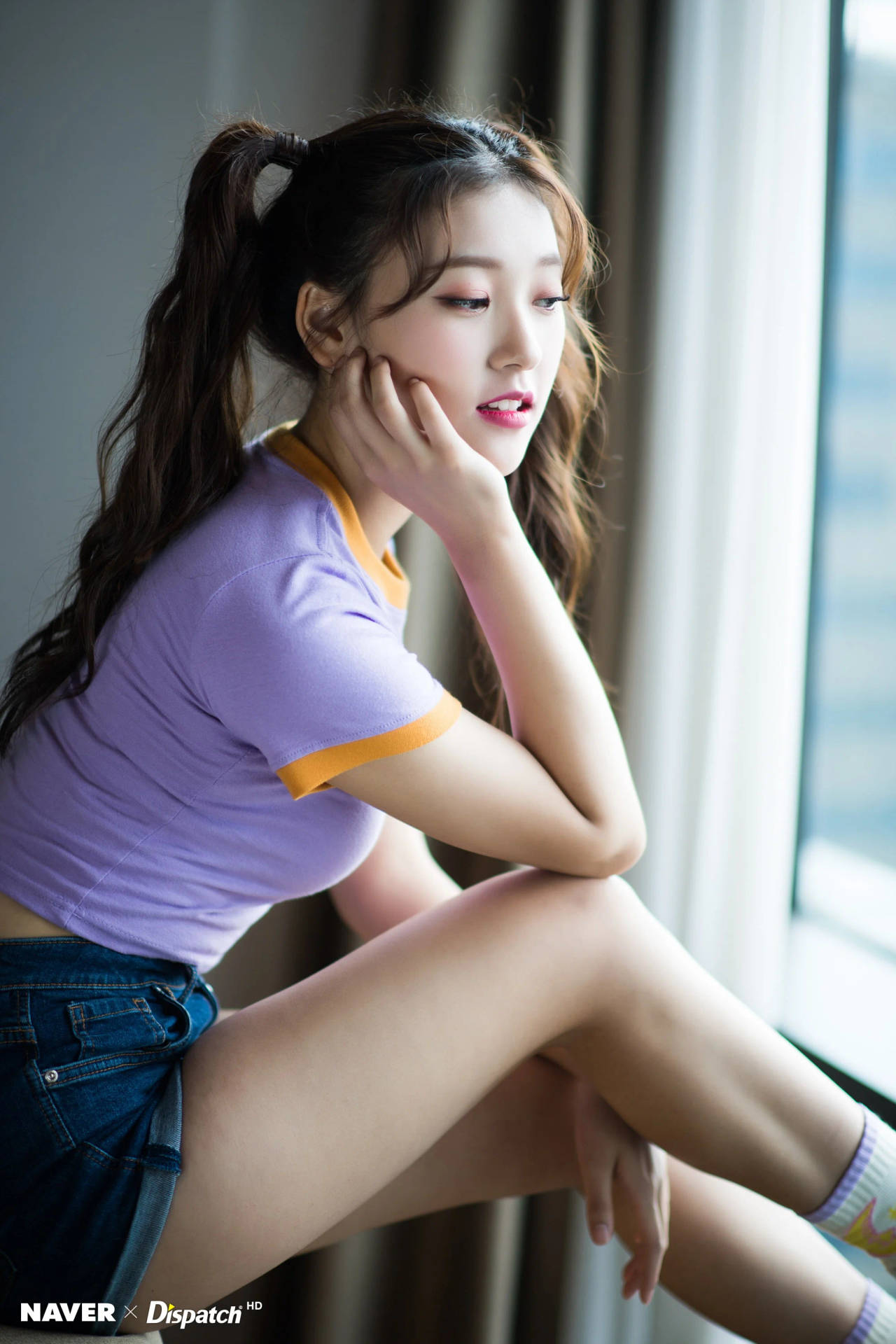 紮辮子的年輕女性穿著紫色襯衫和牛仔短褲坐在窗邊。