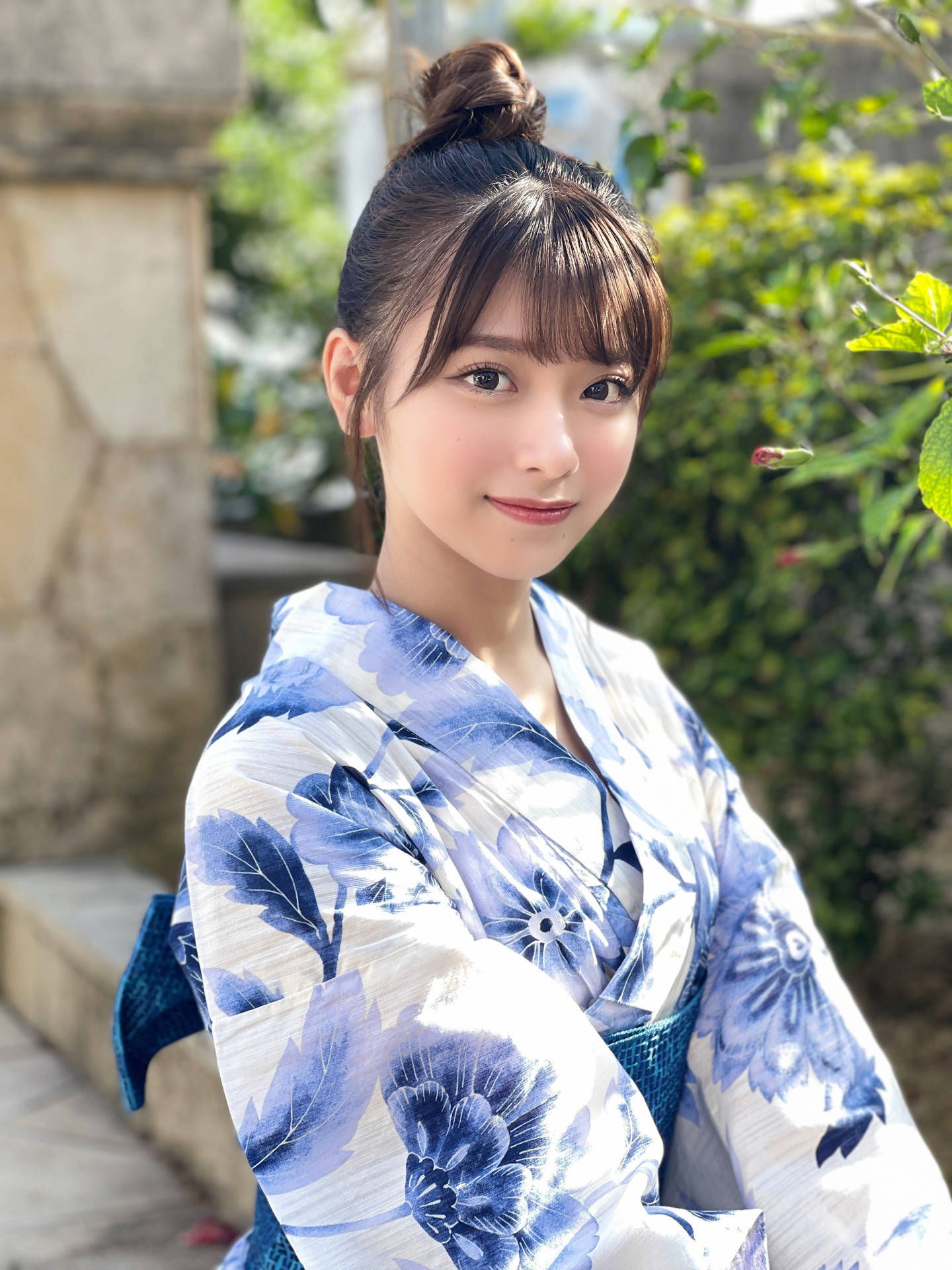 身穿藍色花卉和服的年輕女性在花園中微笑，背景有石牆。