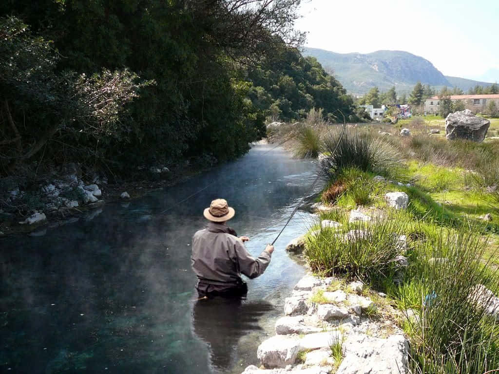 一名男子在山間溪流釣魚，戴著帽子，在清澈的水中涉水。